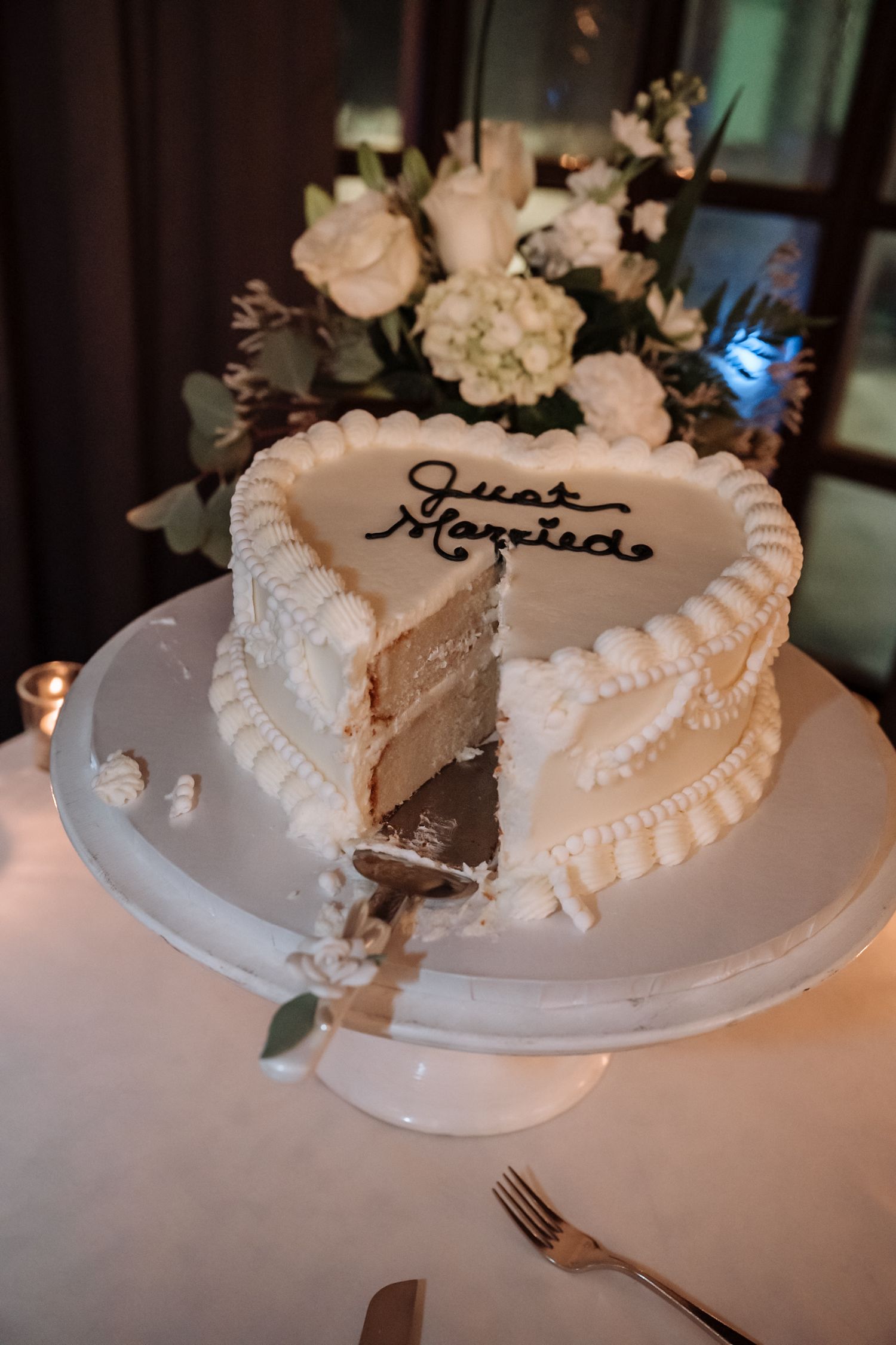 Heart-shaped Just Married wedding cake with white frosting on a cake stand, with a slice removed revealing layers inside.
