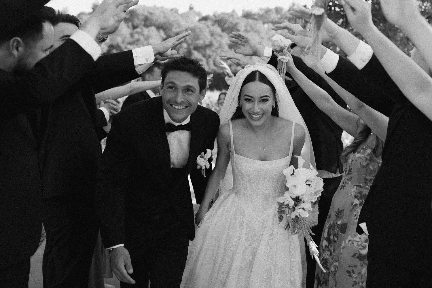 Black and white wedding photo of smiling bride and groom walking through a tunnel of raised hands from guests.