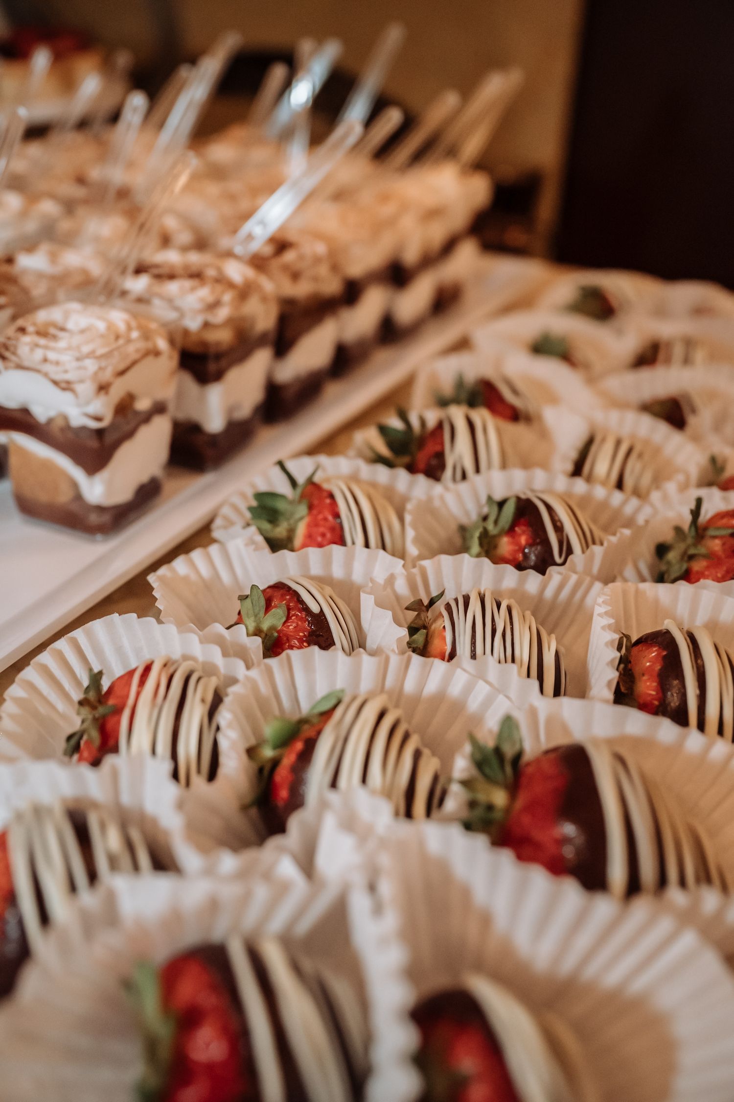 Chocolate-dipped strawberries in paper cups and layered dessert cups displayed on a dessert table.