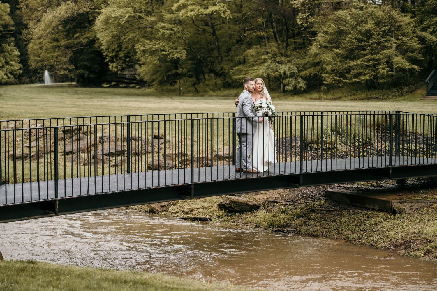 Bride and groom embracing on a bridge over a stream in a lush green park on their wedding day.