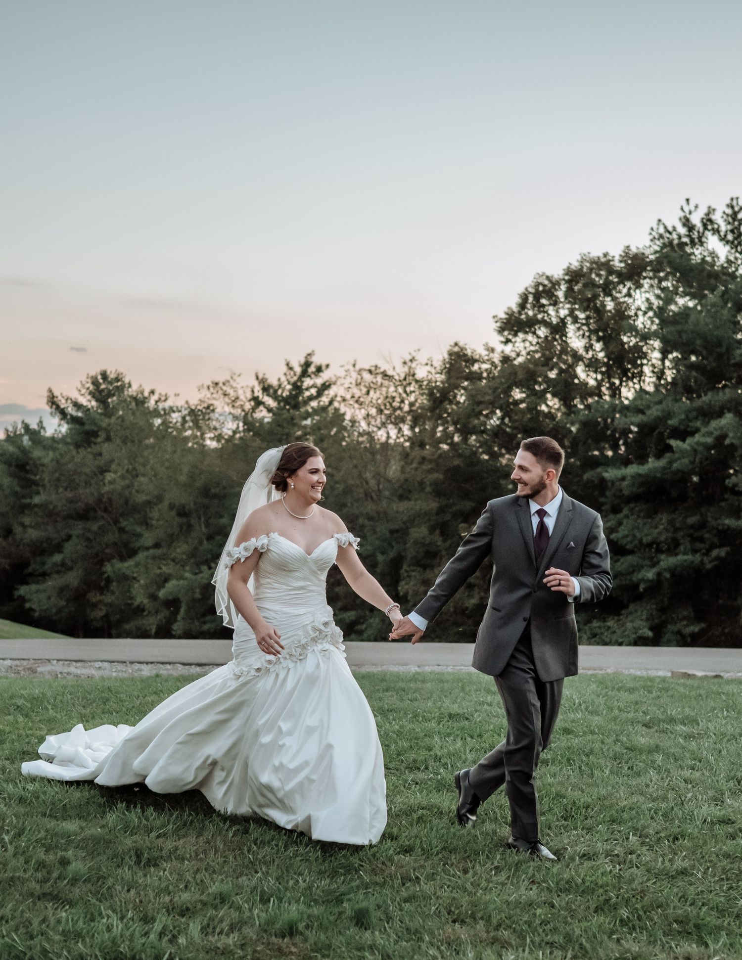 A bride and groom hold hands walking on green grass at dusk, with trees and pastel sky behind them.