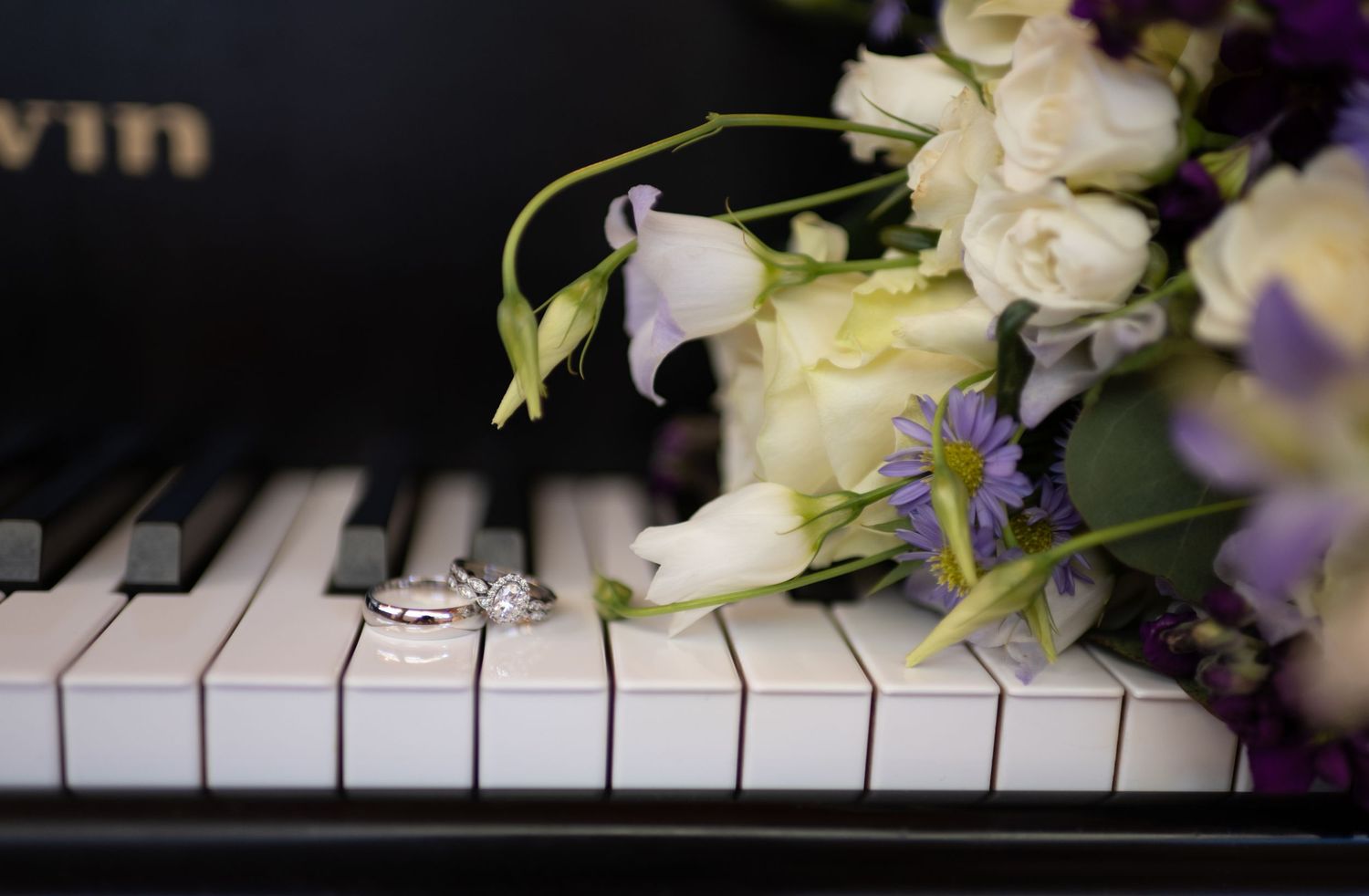 Wedding rings and bridal bouquet with white roses and purple flowers resting on black and white piano keys.