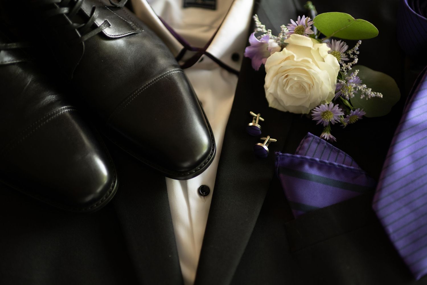 Black dress shoes and a white rose boutonniere with purple flowers on a dark tuxedo jacket with purple tie.
