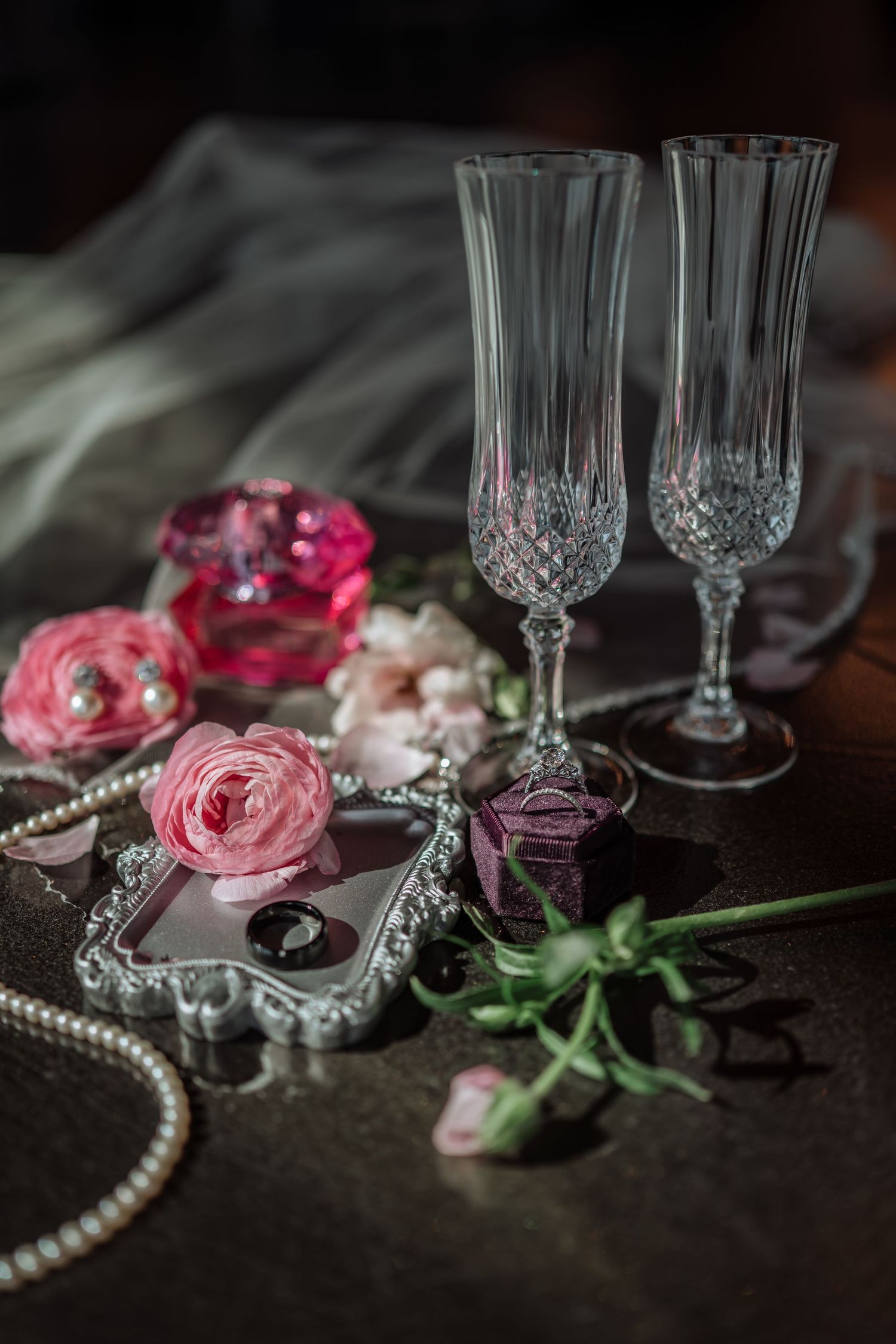 Elegant wedding flat lay with crystal champagne flutes, pink ranunculus flowers, pearl necklace, and rings on a dark surface.
