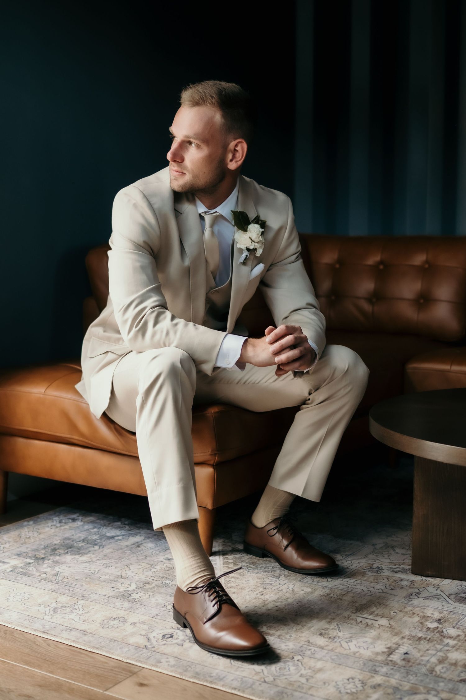 Groom in beige suit with boutonniere sits on brown leather sofa, gazing away in elegant dark-walled room.