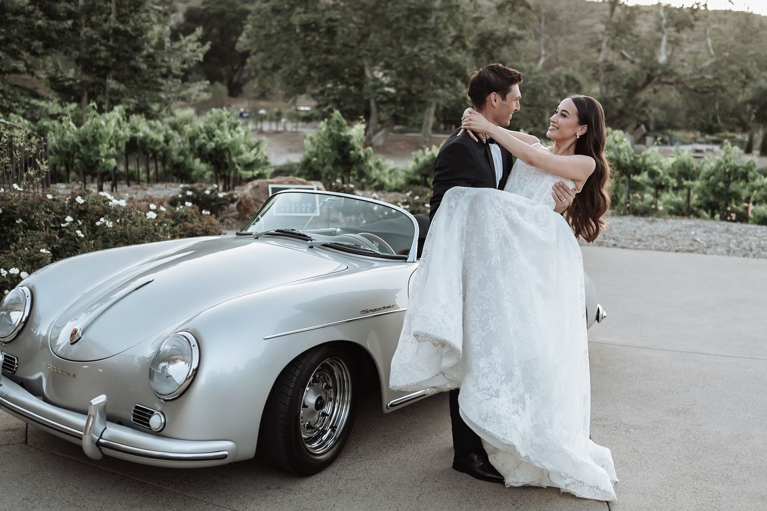 Bride and groom posing romantically beside a vintage silver Porsche 356 convertible in a garden setting.