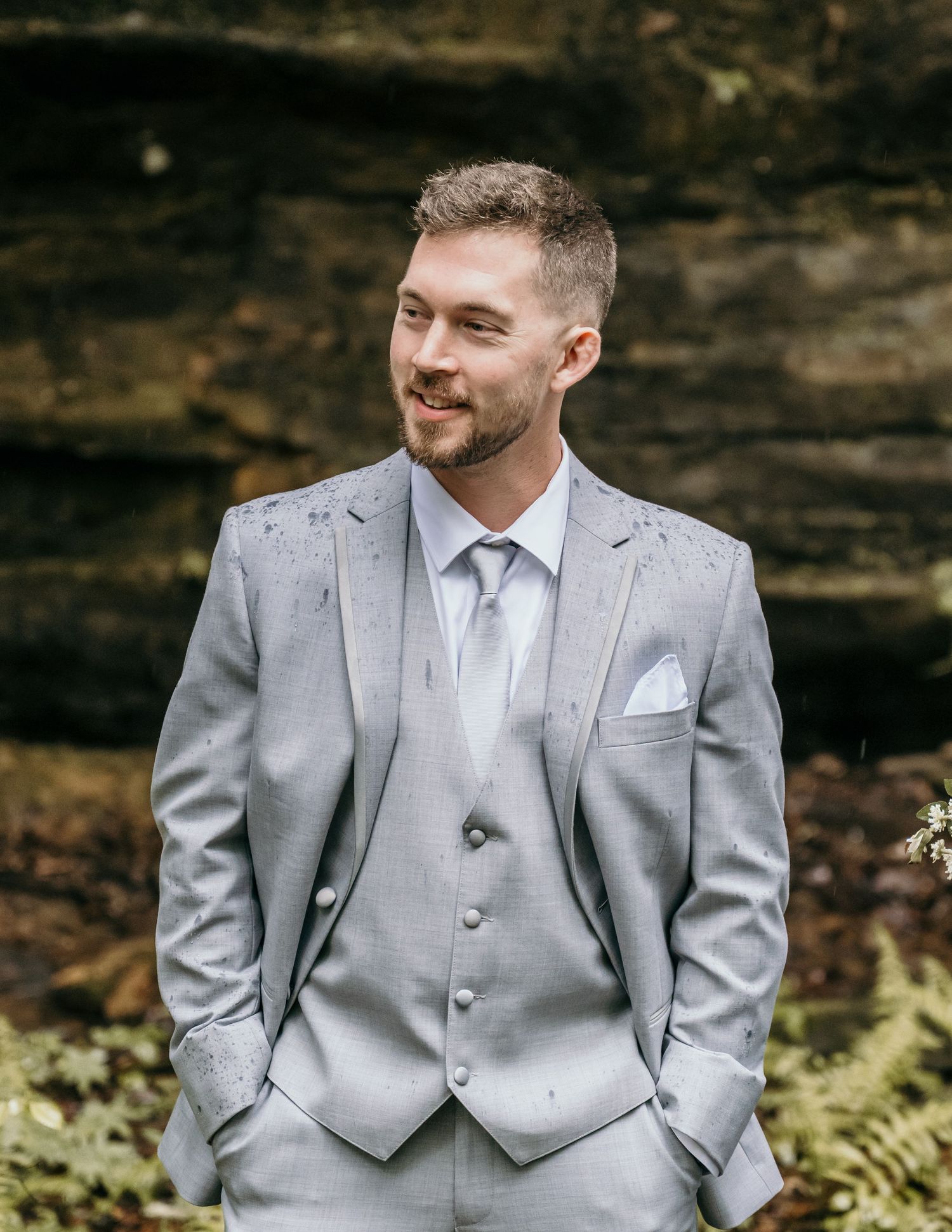 A smiling groom in a light gray three-piece suit with white pocket square and tie stands outdoors near a stone wall.