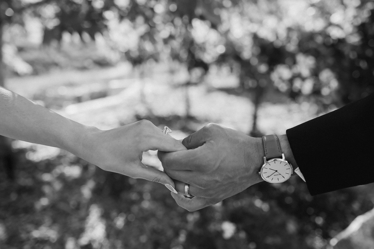 Black and white photo of a couple holding hands, man wearing a watch, with blurred greenery background.