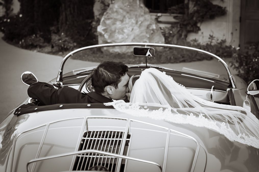 Black and white photo of a bride and groom sharing a kiss in a vintage convertible car on their wedding day.