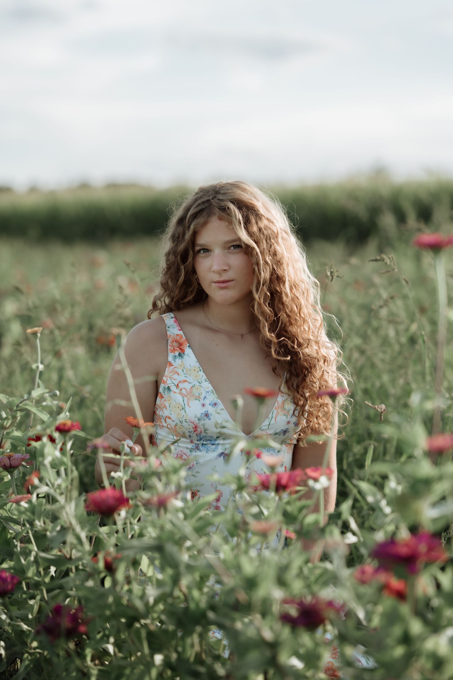 Young woman with curly hair in floral dress sitting among wildflowers in a lush green field.