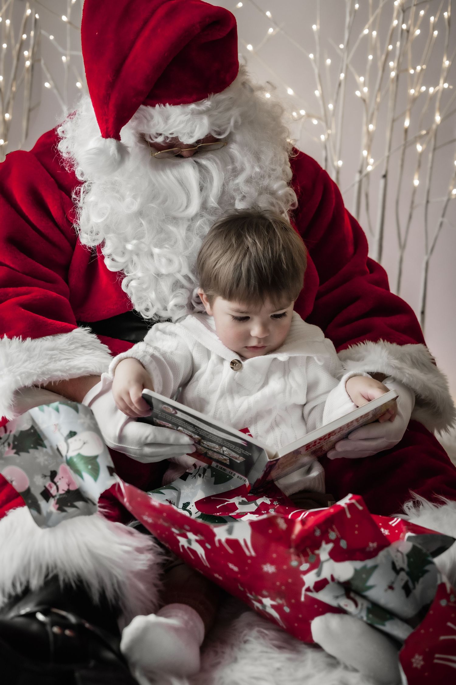 Toddler in white pajamas sits on Santa Claus lap reading a book surrounded by festive Christmas decorations.