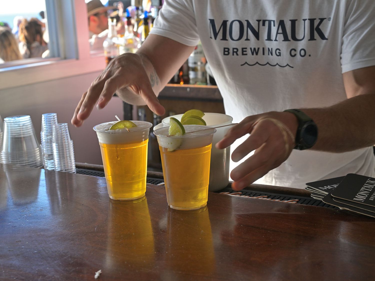 Bartender at Montauk Brewing Co. serving two plastic cups of beer garnished with lime wedges on a wooden bar.