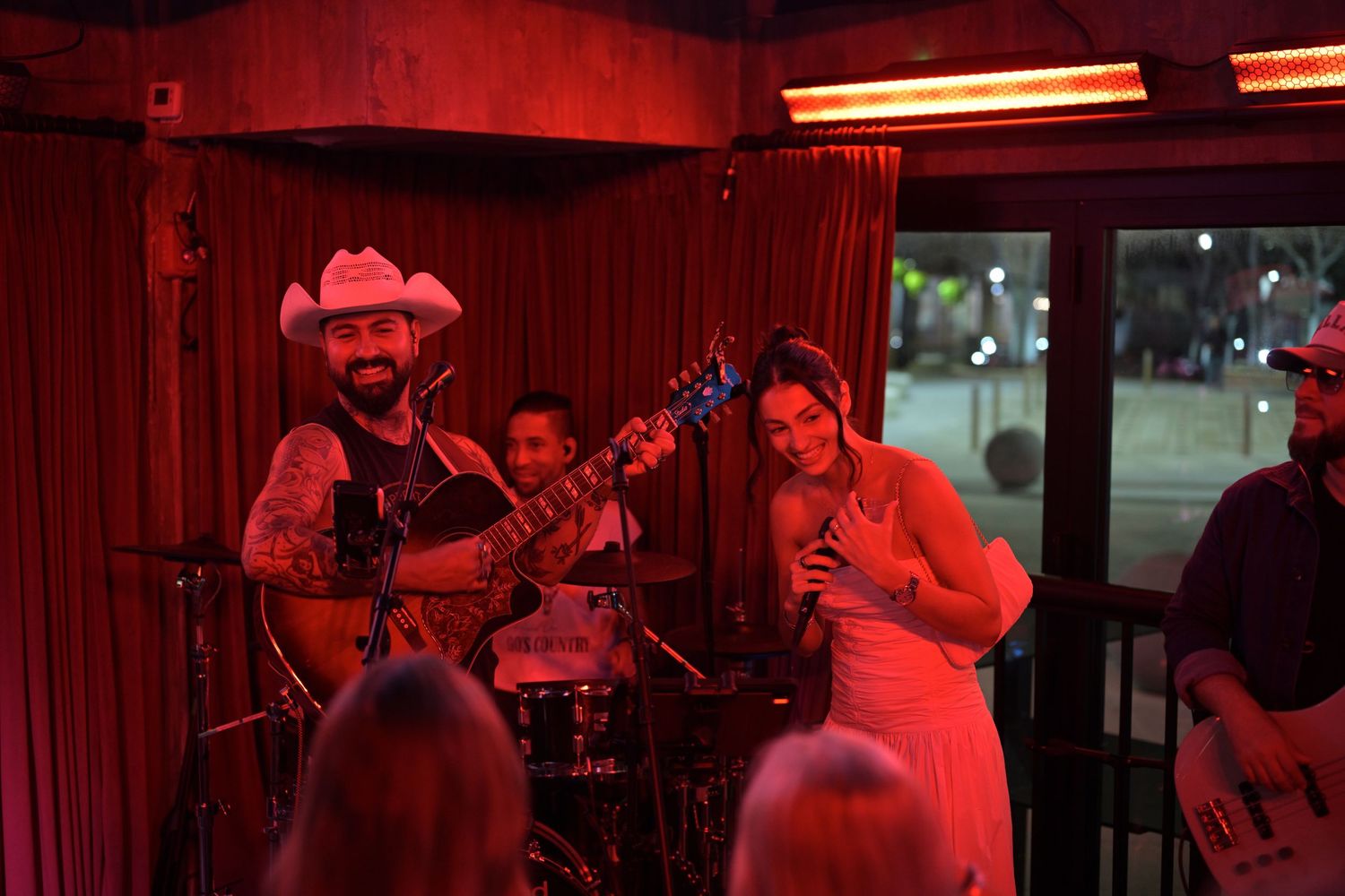 Country band performing on a red-lit stage with a bearded guitarist in a cowboy hat and female vocalist smiling at crowd.