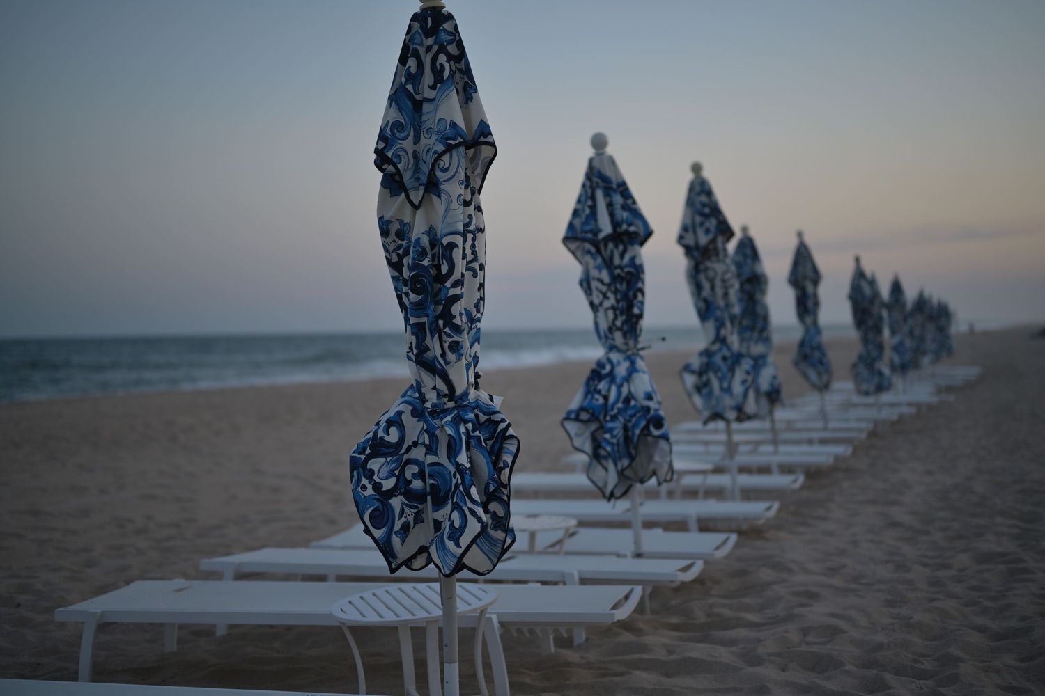 Row of folded blue and white patterned beach umbrellas with lounge chairs on a sandy beach at dusk.