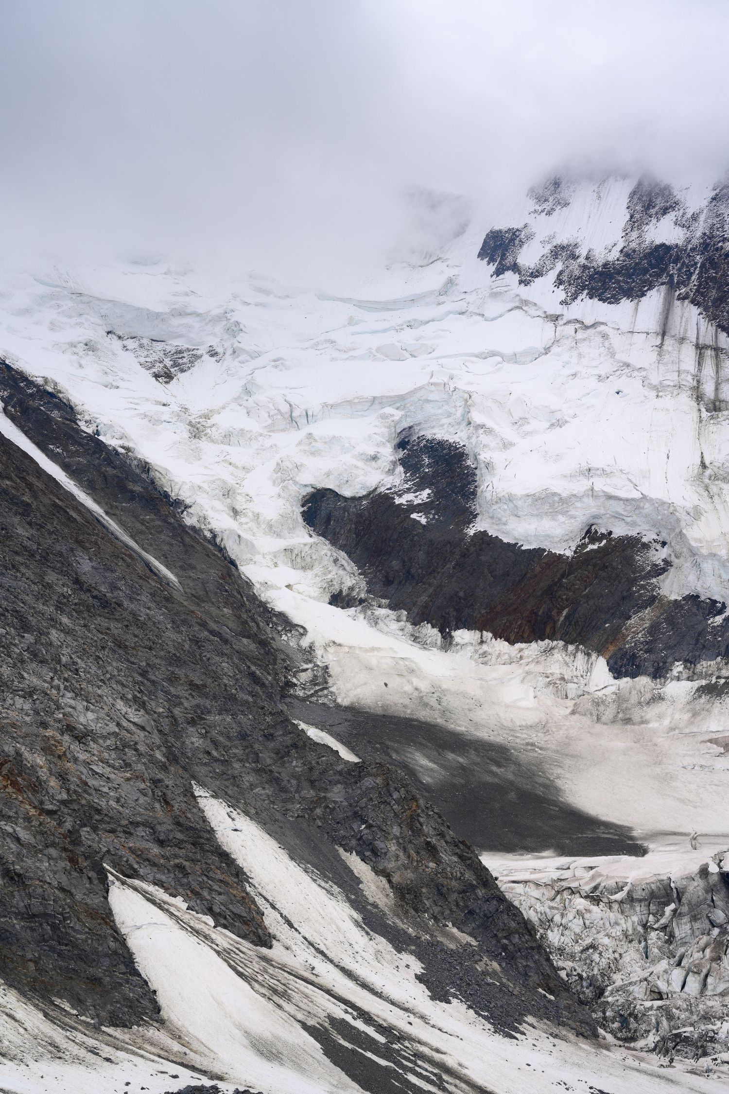 Dramatic alpine glacier with snow and ice flowing between rocky mountain ridges under a cloudy overcast sky.