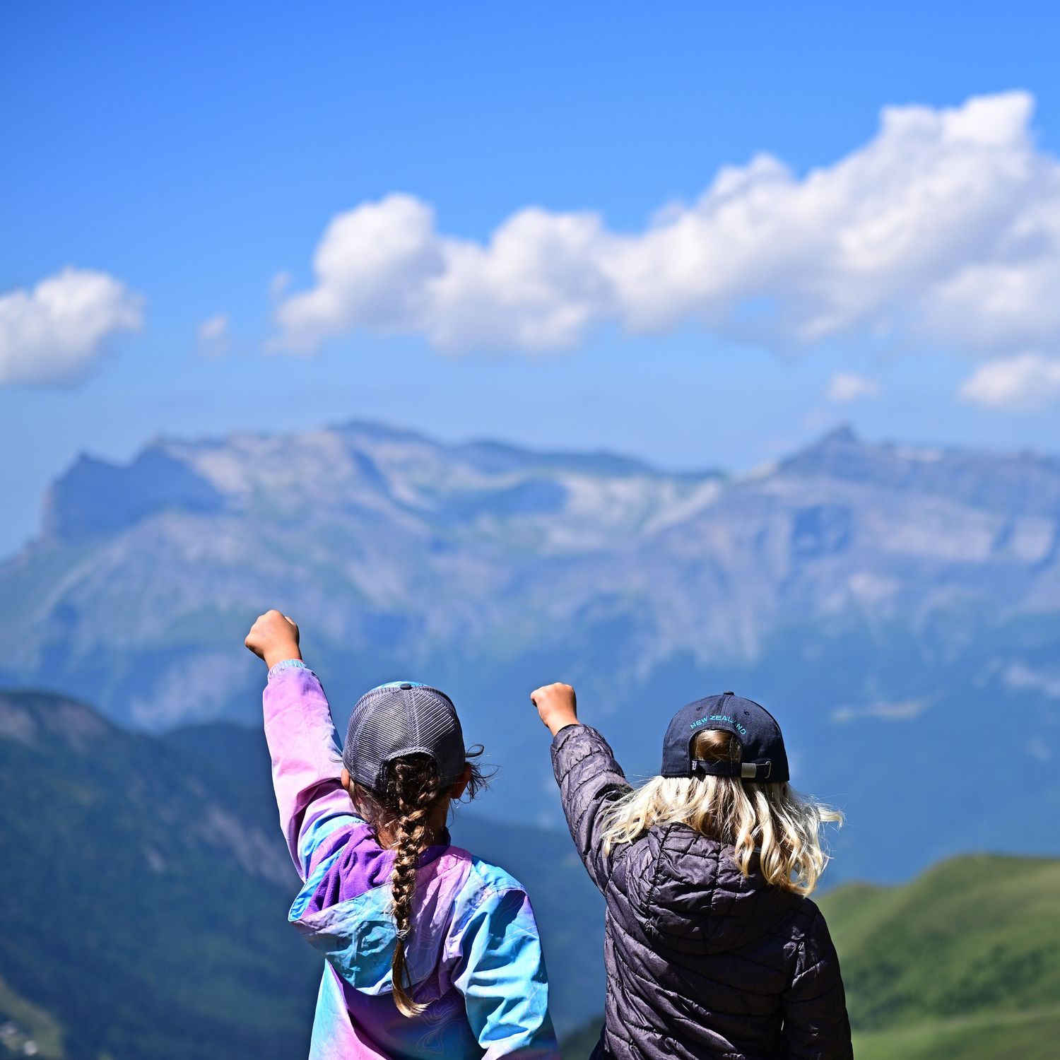 Two children celebrating with raised fists at a mountain summit overlooking scenic alpine peaks.
