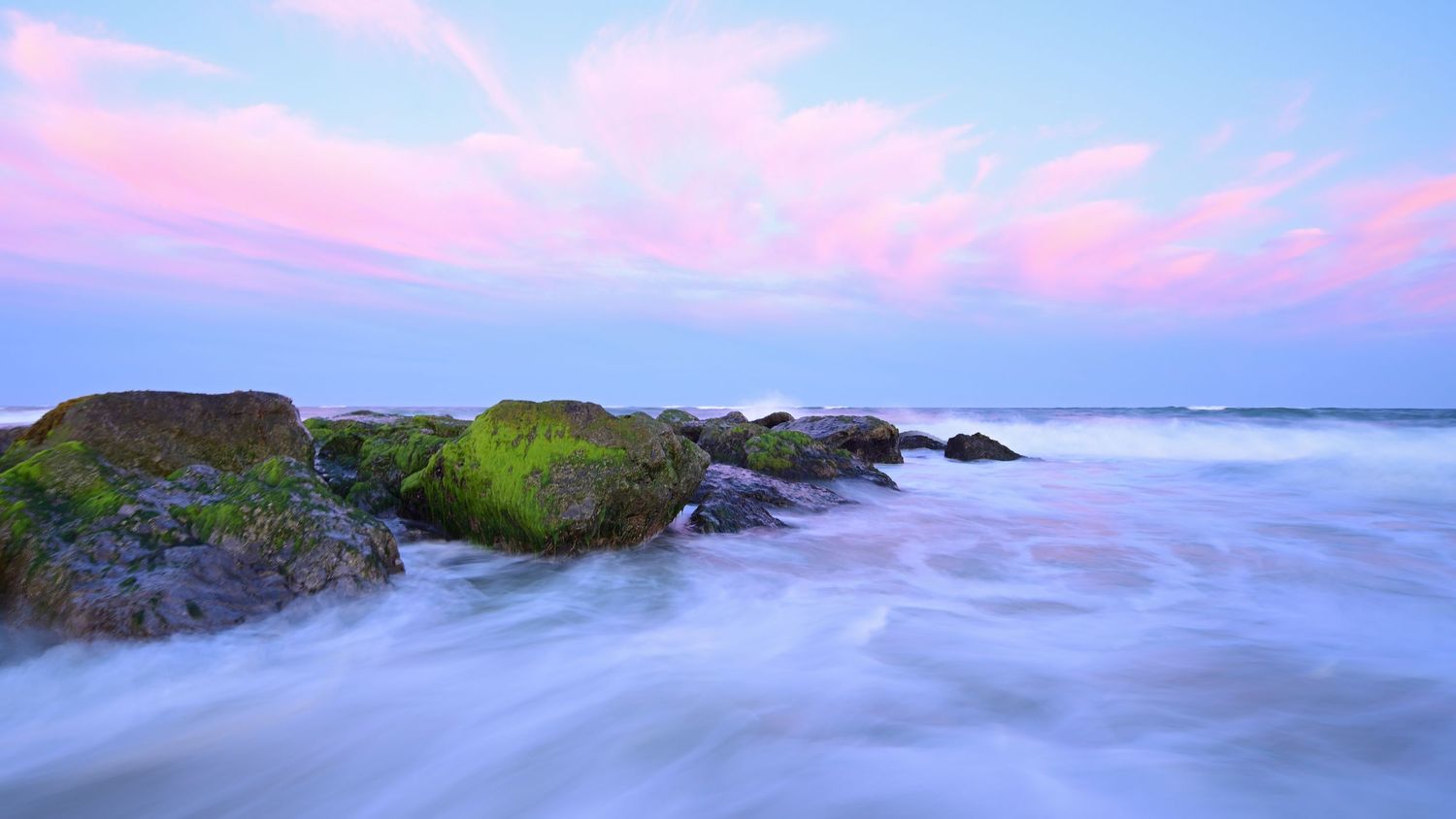 Serene long exposure seascape featuring moss-covered rocks and smooth flowing ocean water under a pastel pink sky.