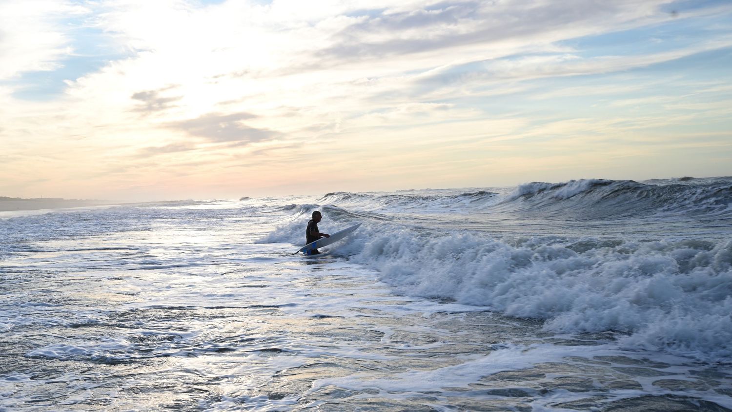 Lone surfer riding waves at sunset on a dramatic cloudy beach with crashing surf.