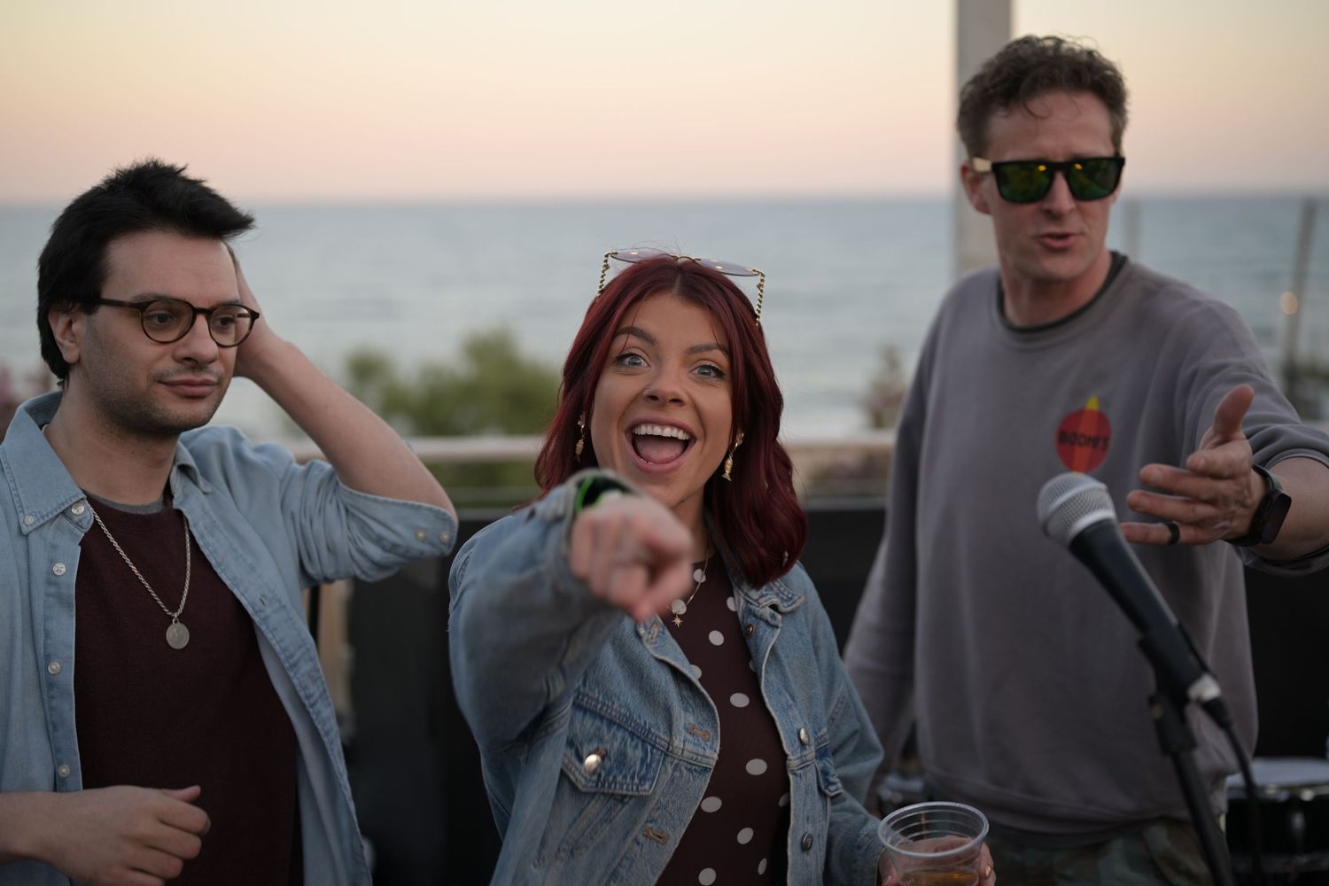 Three people laughing and having fun at an outdoor beachside event at sunset with a microphone nearby.