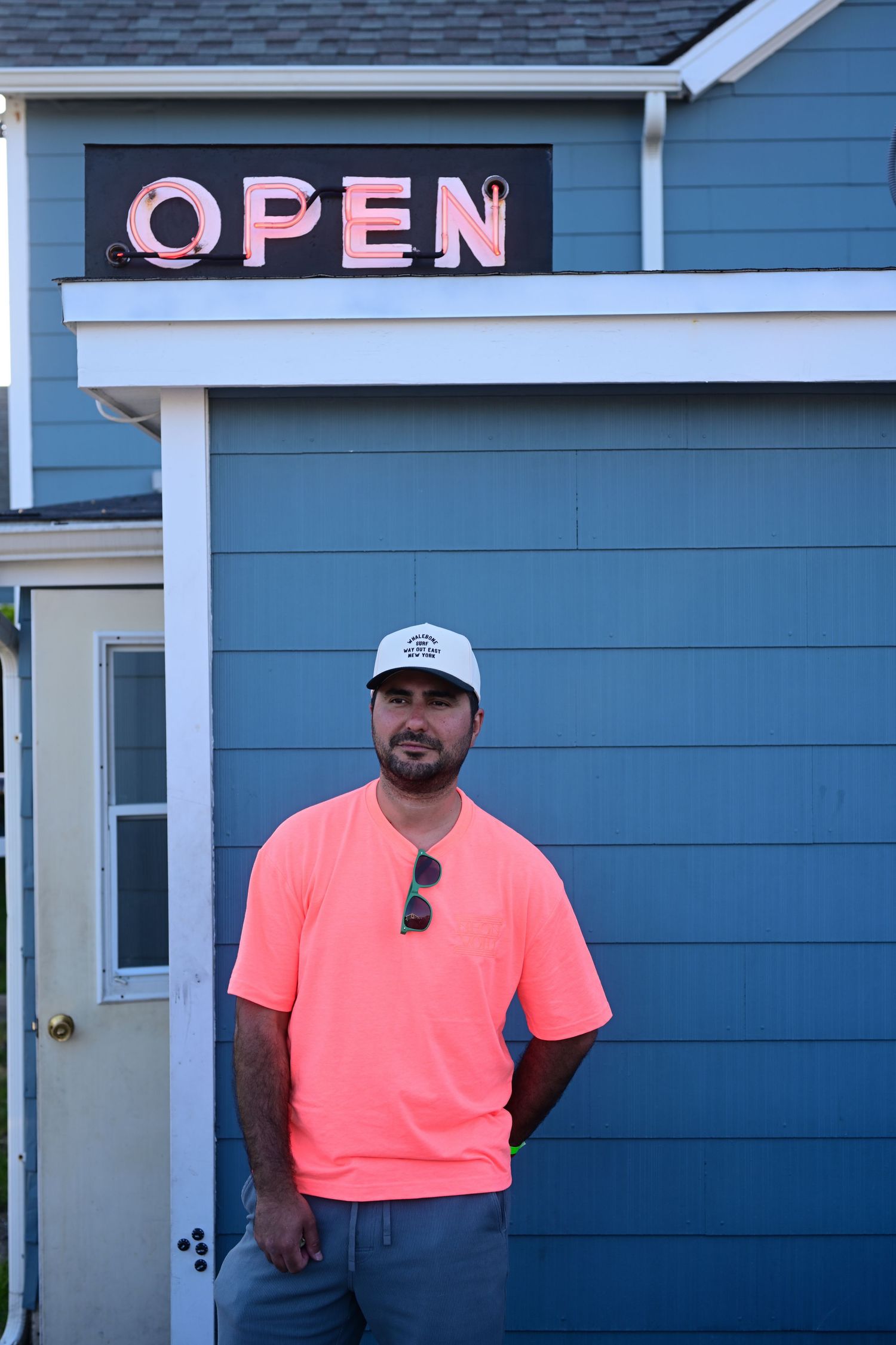 Man in pink t-shirt and white cap standing under an Open sign outside a blue wooden building.