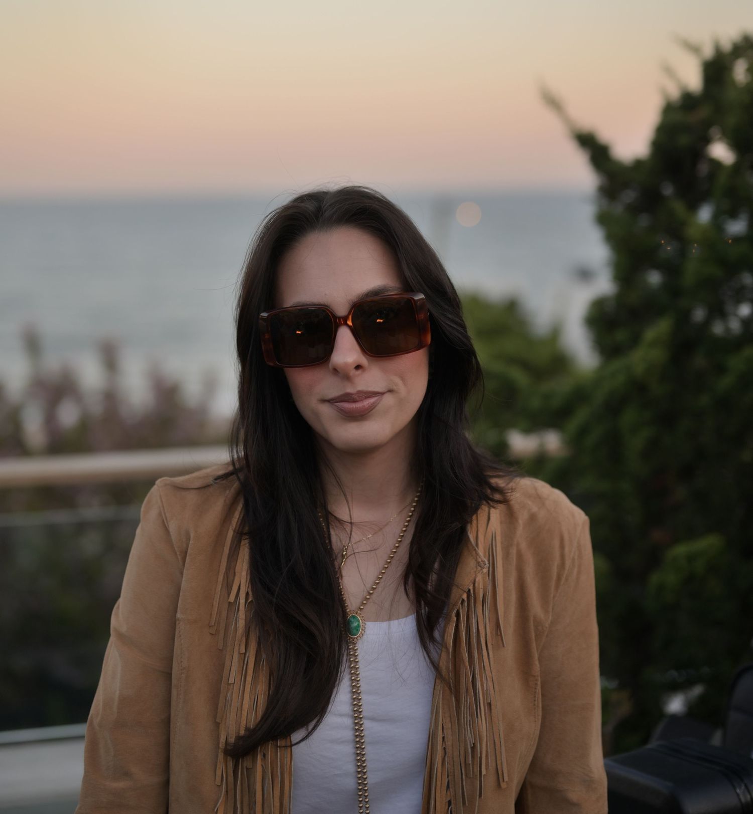 Young woman with long dark hair wearing sunglasses and a tan fringe jacket poses at sunset near the ocean.
