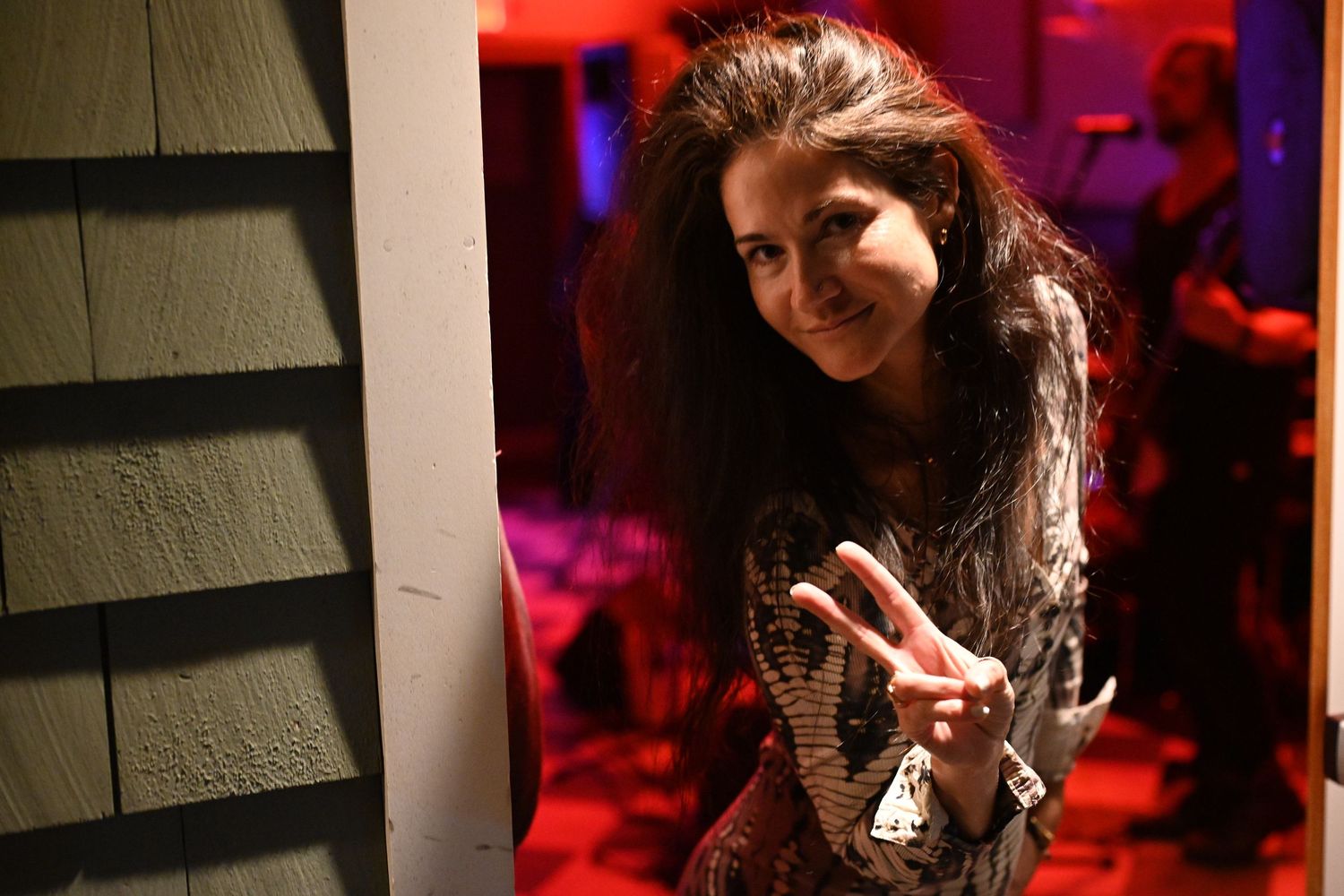 Woman with long dark hair making a peace sign while peeking around a corner in red-lit party setting.