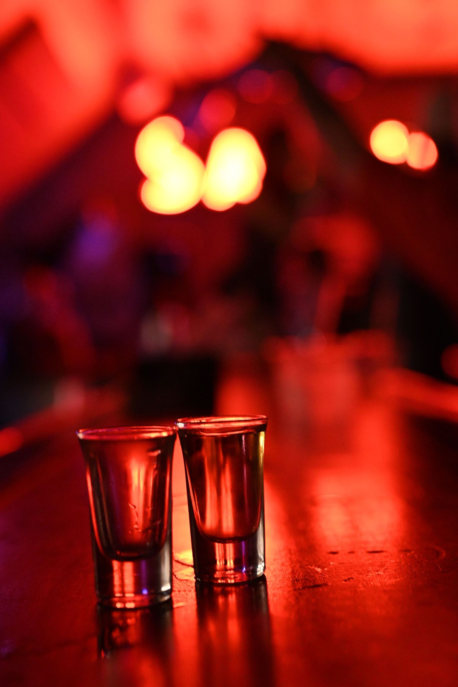 Two shot glasses on a wooden bar counter with blurred red bokeh lights in the background.