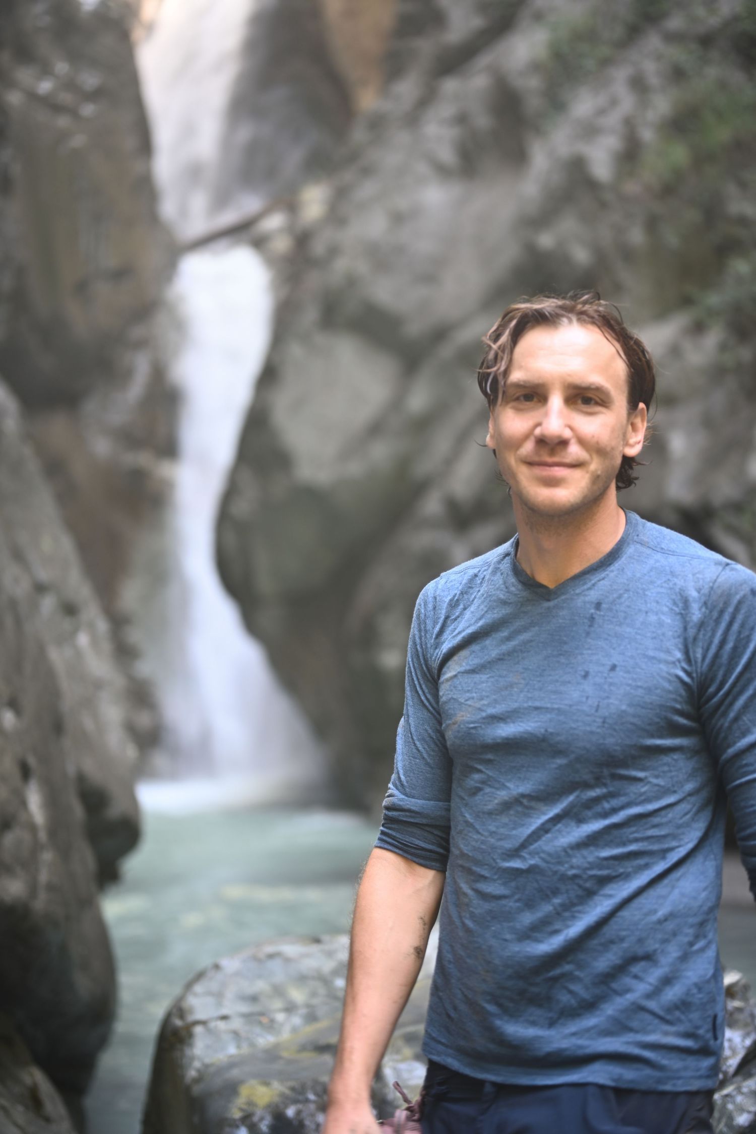 Man smiling in front of a waterfall cascading between rocky canyon walls, wearing a blue t-shirt outdoors.