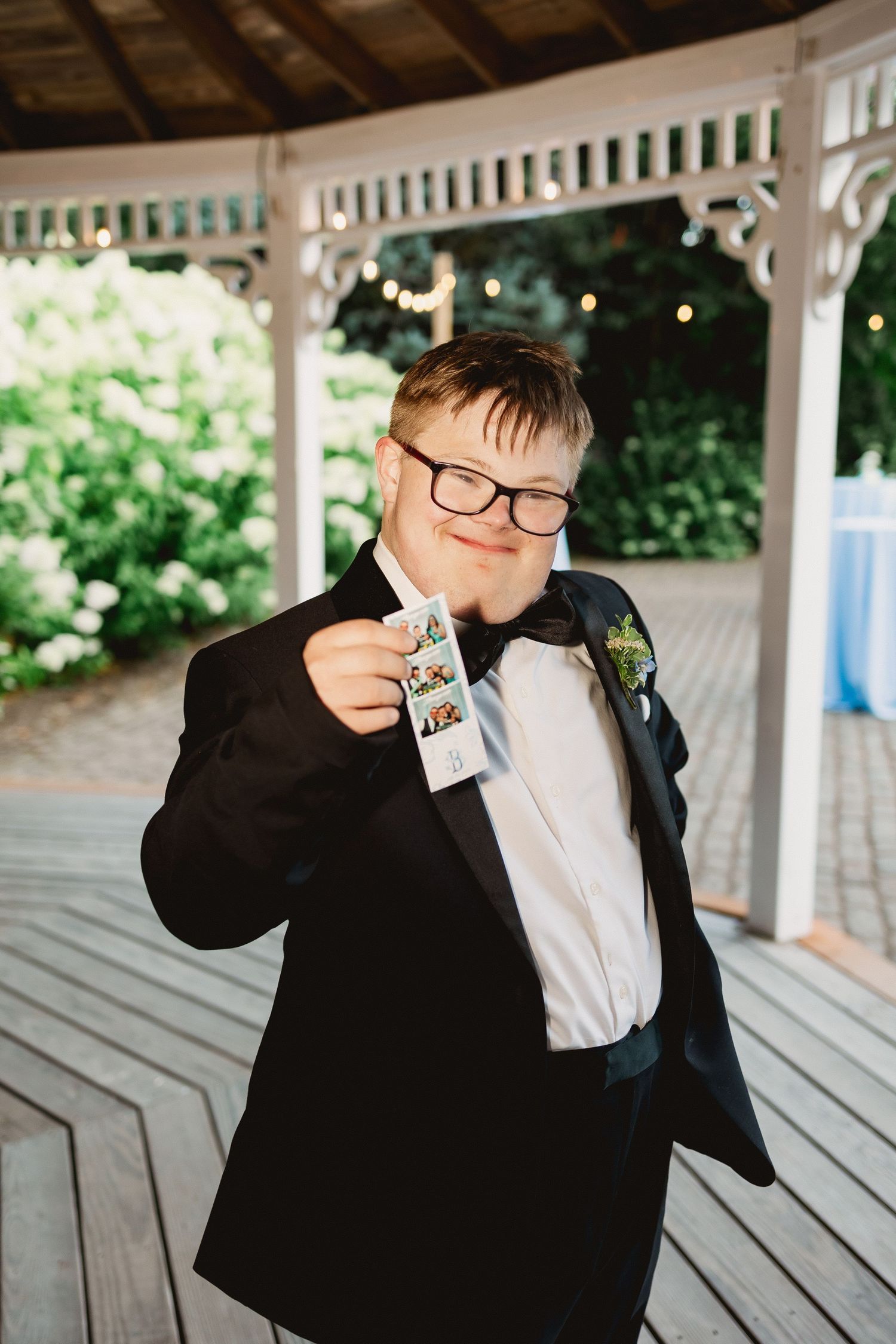 A person in a black suit and bowtie takes a drink from a glass bottle on a covered wooden deck.