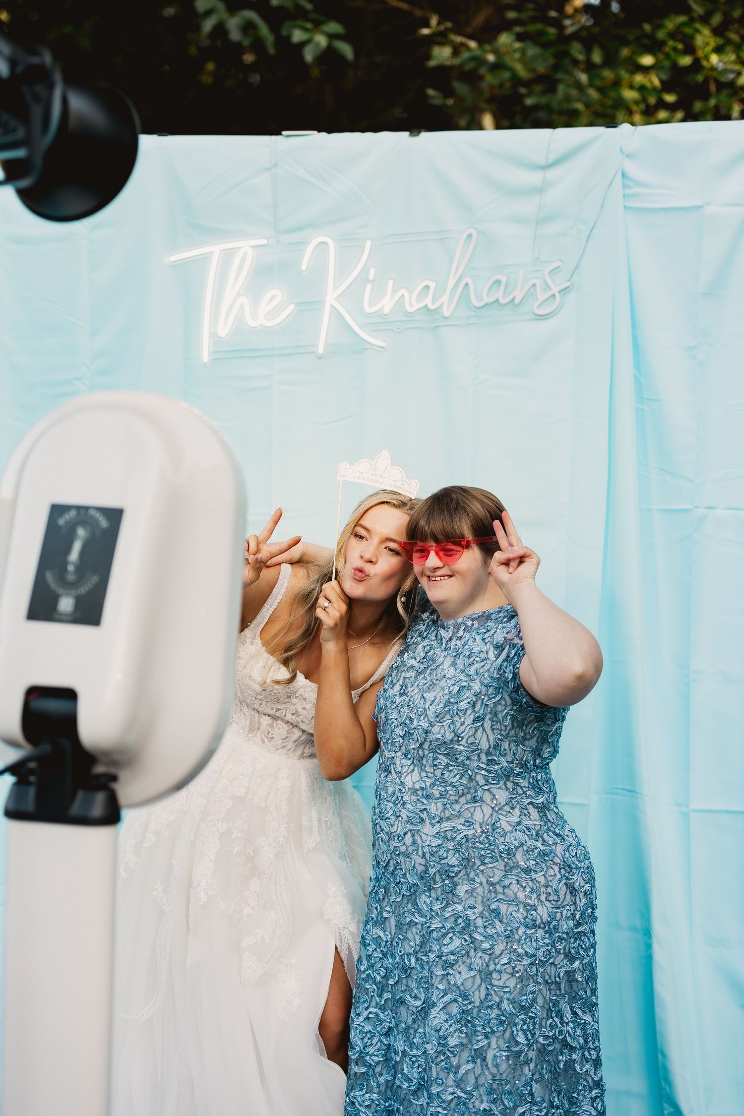 Two people pose playfully in front of a light blue backdrop with neon sign that says 'The Kinchans' at a photo booth.