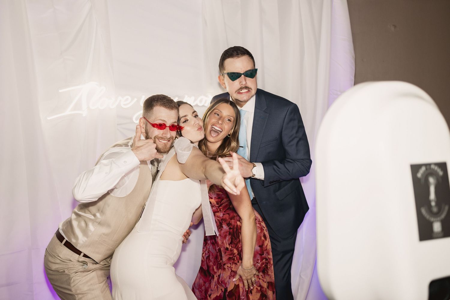 Three people pose together at a party wearing sunglasses and festive attire in front of a white backdrop.
