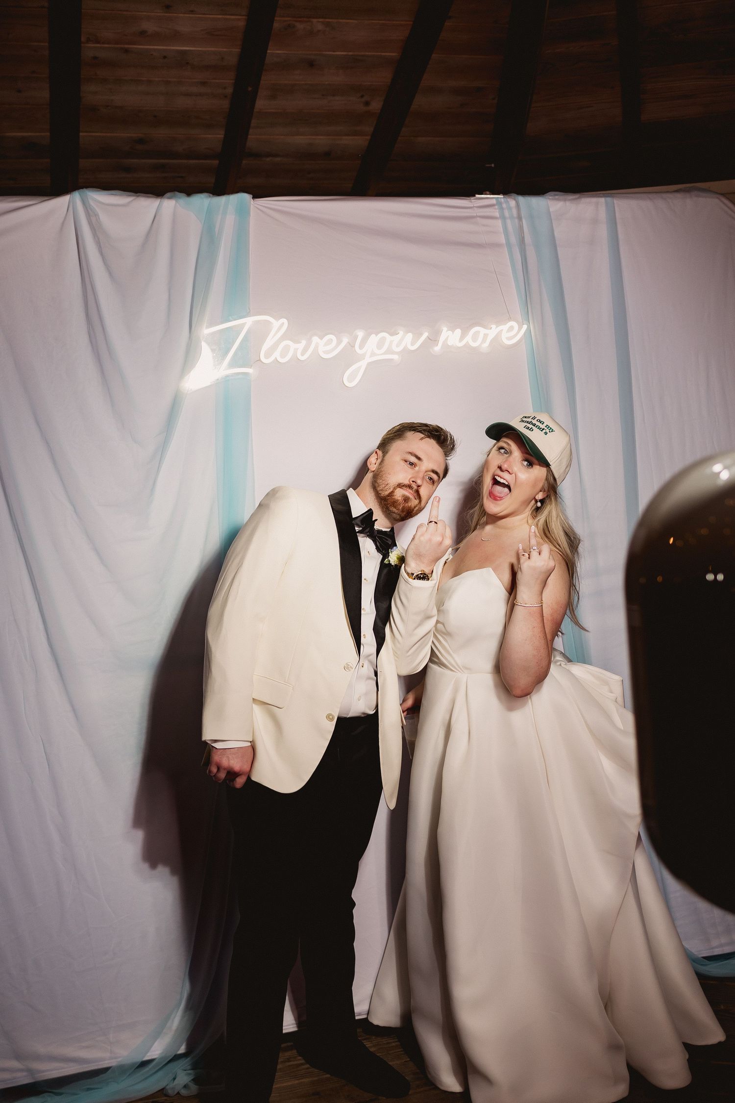 Newlywed couple laughing and posing playfully in front of white backdrop at wedding reception.