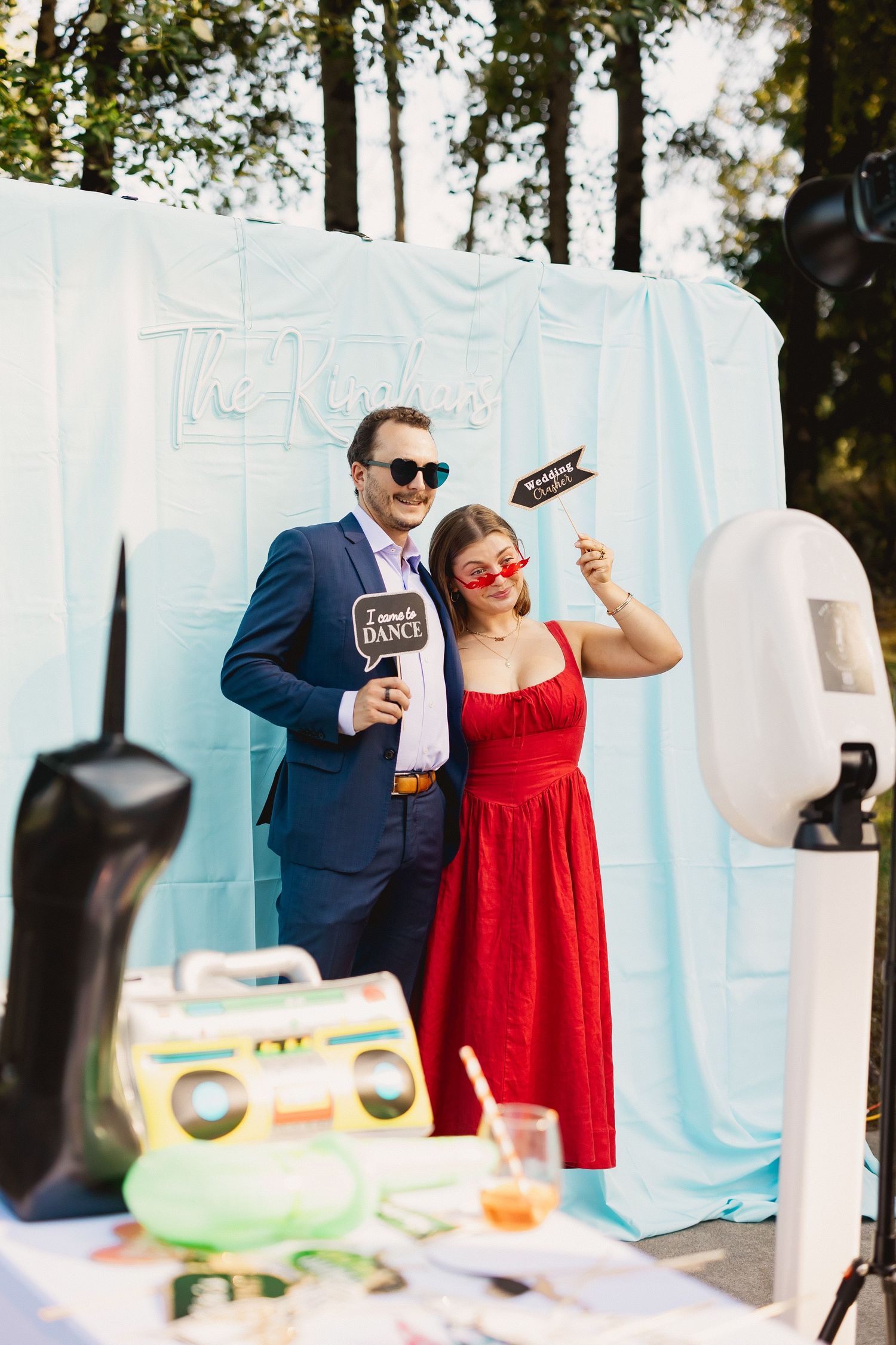 A couple in formal attire poses playfully with props at an outdoor photo booth with white backdrop and sunlit trees.