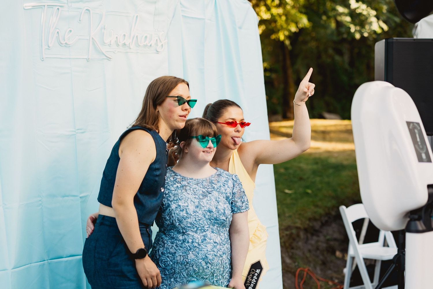A wedding party enjoys outdoor festivities with white chairs and sunlit trees in the background.