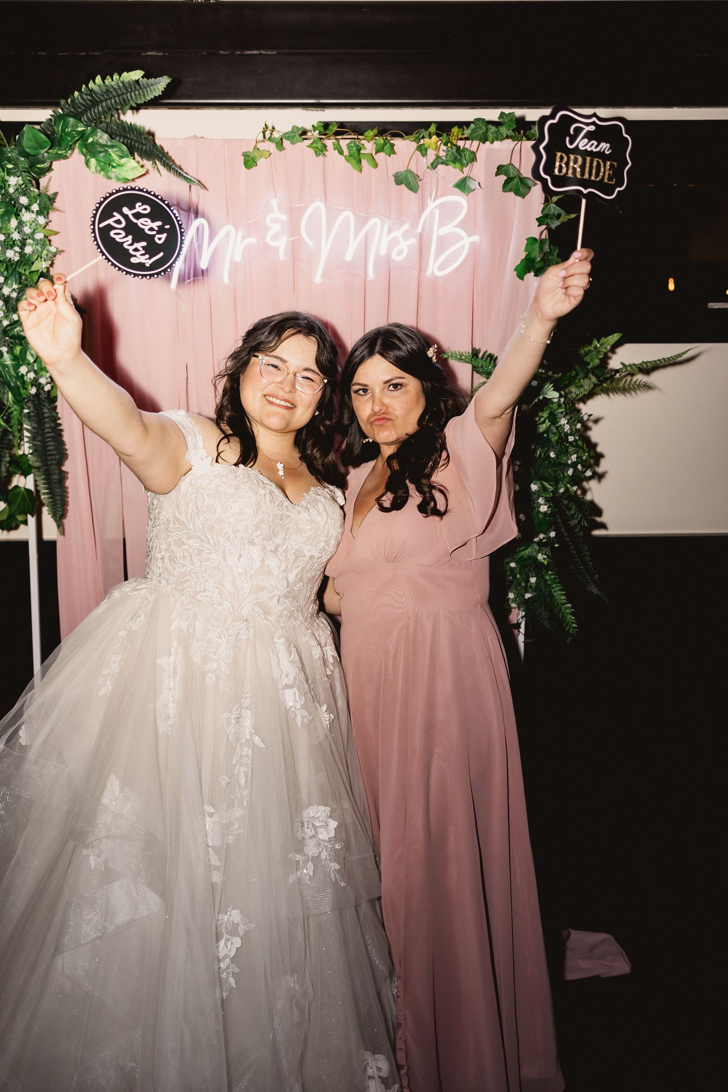 Wedding celebration with bride in white gown and bridesmaid in dusty rose dress posing together at photo booth.