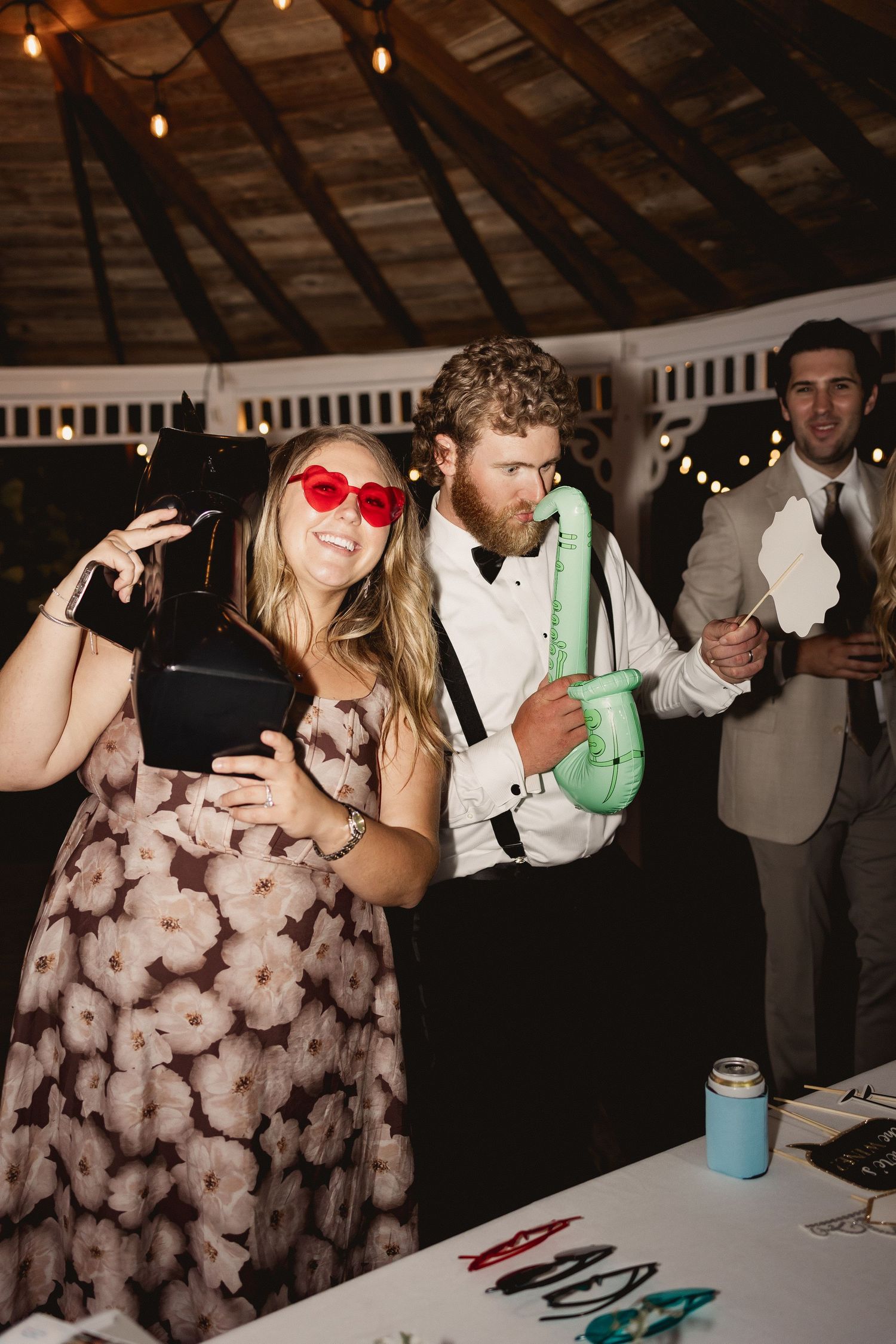 Wedding guests enjoy dancing and posing with props at a rustic barn reception with string lights overhead.