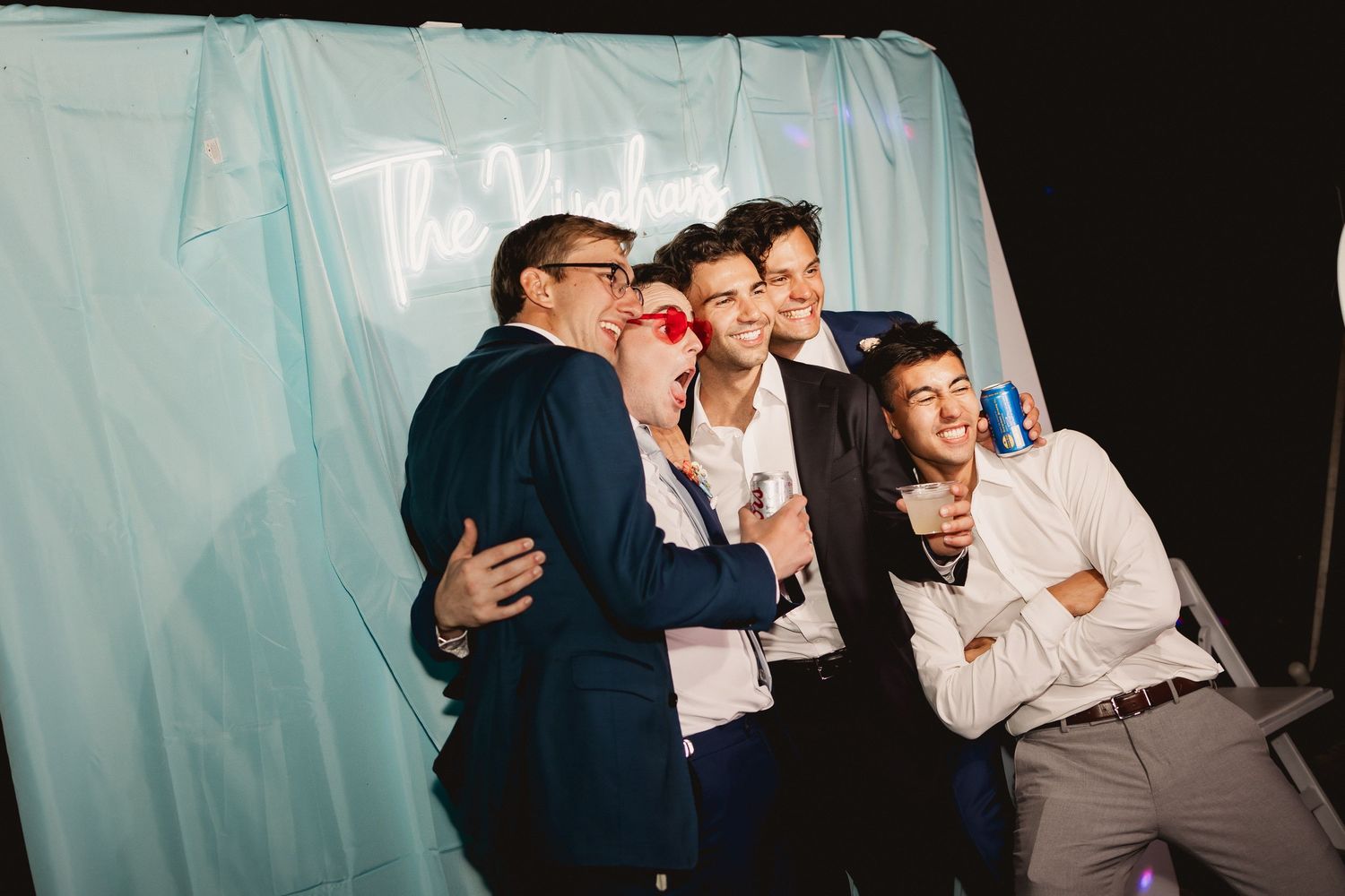 A group of people laughing and embracing at a party with light blue curtains in the background.