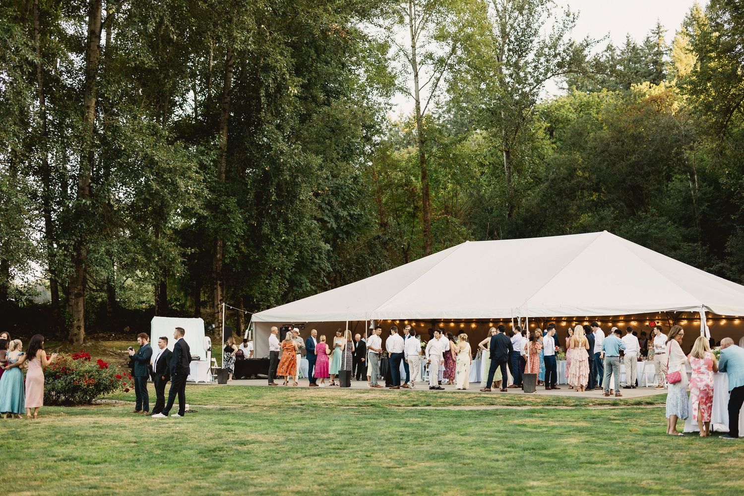 Outdoor wedding reception under a white tent on a green lawn surrounded by tall trees at dusk.