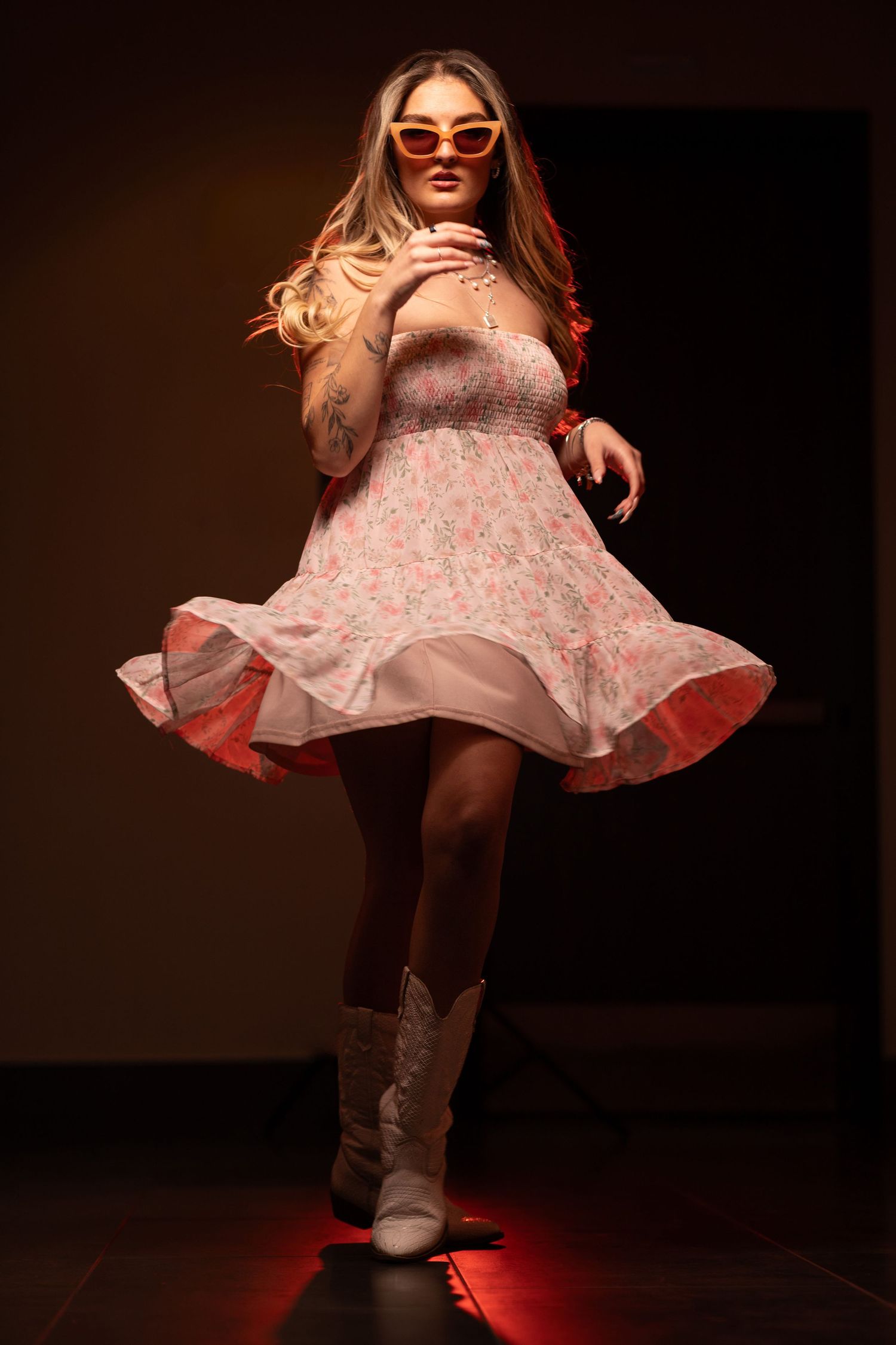 Dynamic indoor portrait of a stylish woman in a floral sundress and white cowboy boots, captured mid-twirl with dramatic spotlighting