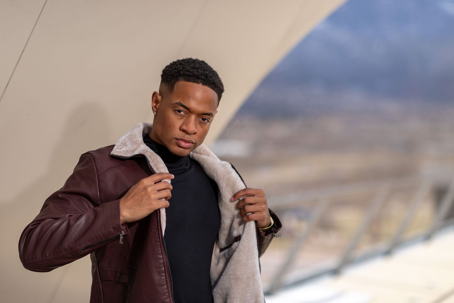 Confident man wearing a maroon leather jacket with shearling collar, leaning on a railing with a blurred mountain background