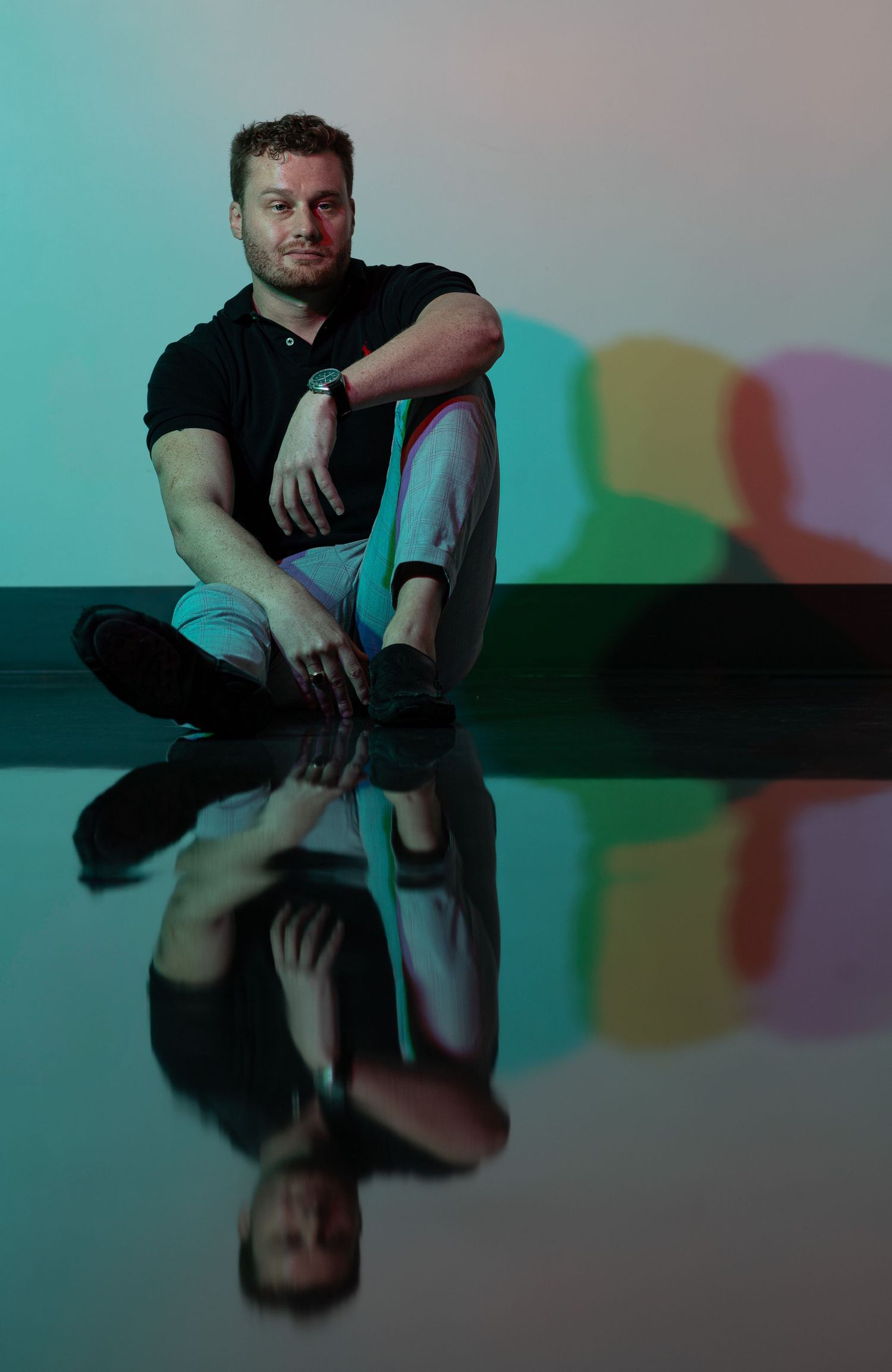 Moody portrait of a seated man reflected on a glossy floor, surrounded by colorful projected shadows. Artistic lighting and reflections