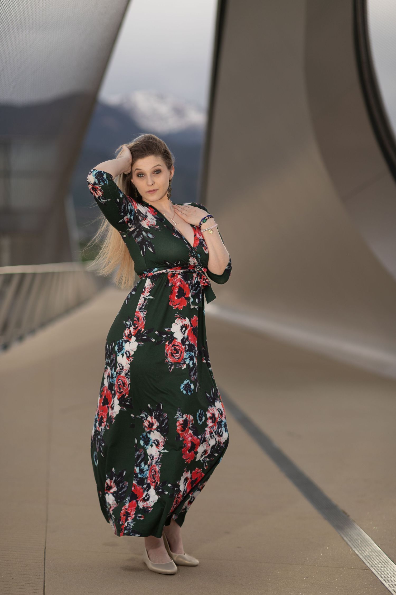 Elegant woman posing on a modern bridge wearing a green floral maxi dress with long blonde hair