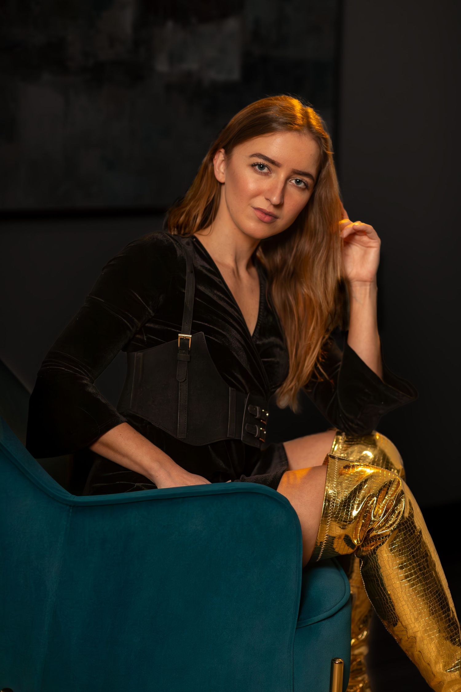 Woman in black velvet dress with gold thigh-high boots sitting on a teal chair in a studio setting.