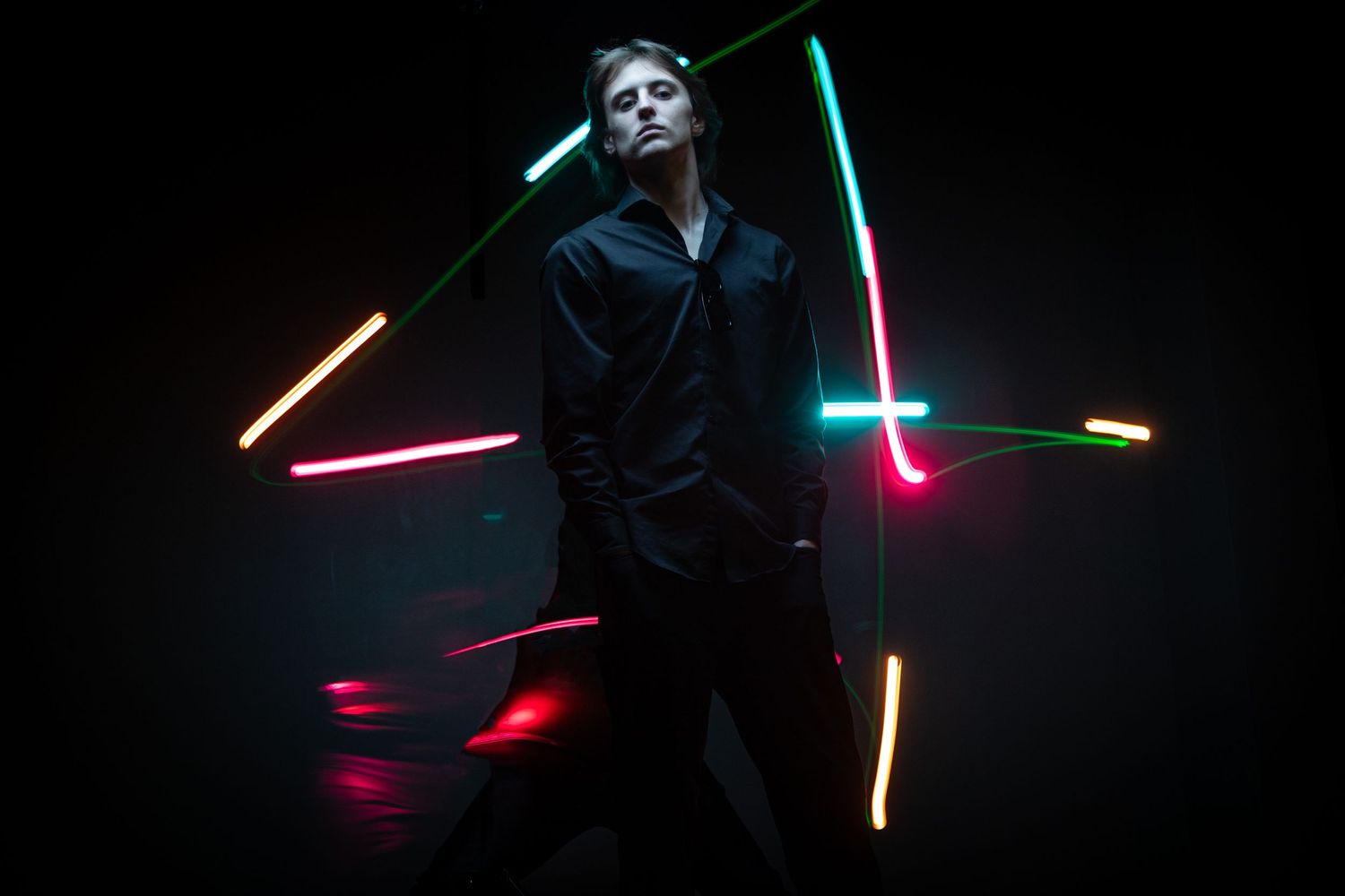 Creative portrait of a man in an all-black outfit illuminated by vibrant neon light trails. Modern studio photography with long exposure