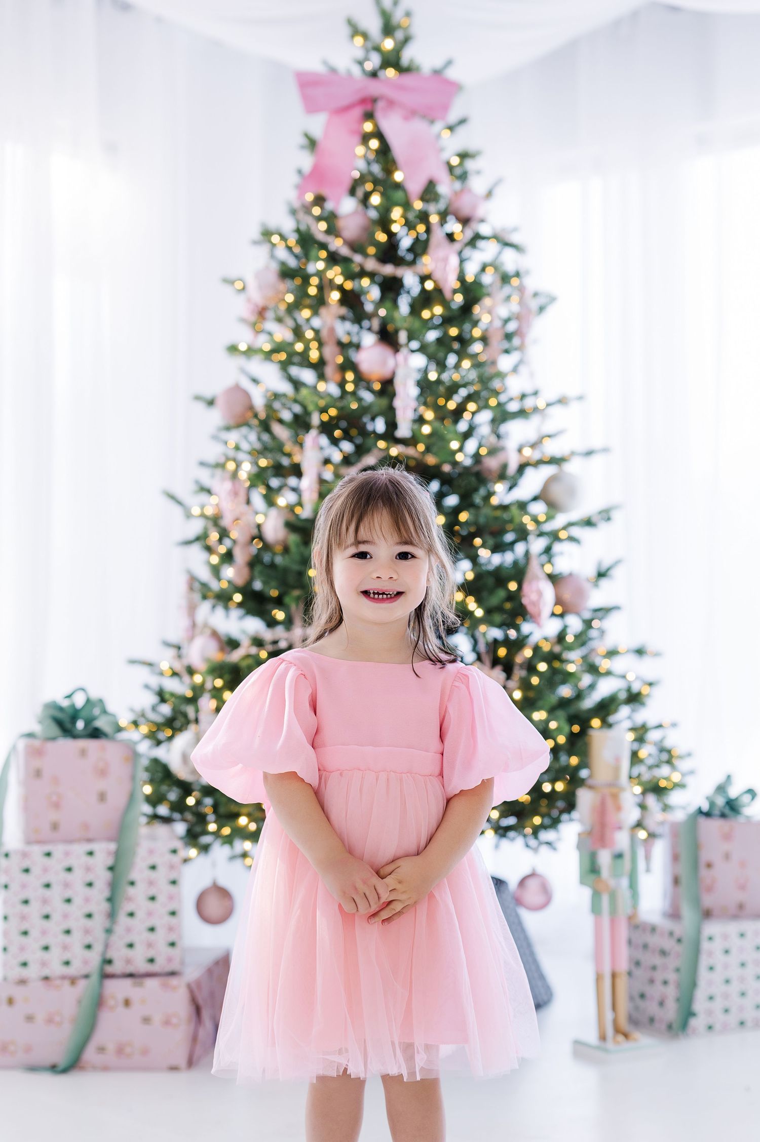 Young girl in pink tulle dress smiling before decorated Christmas tree with pink ornaments and wrapped gifts.