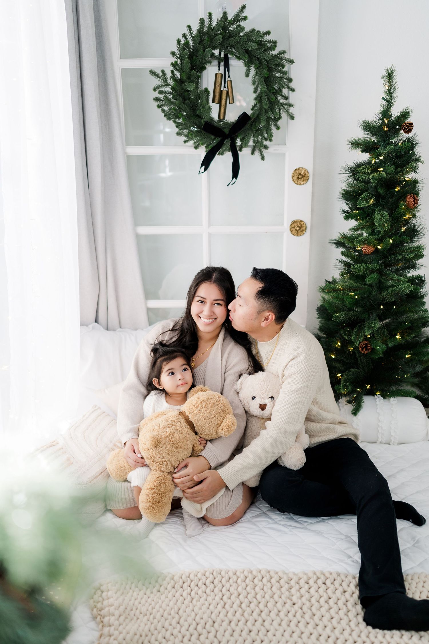 Asian family sitting together on bed with teddy bears near Christmas tree and wreath holiday decor.
