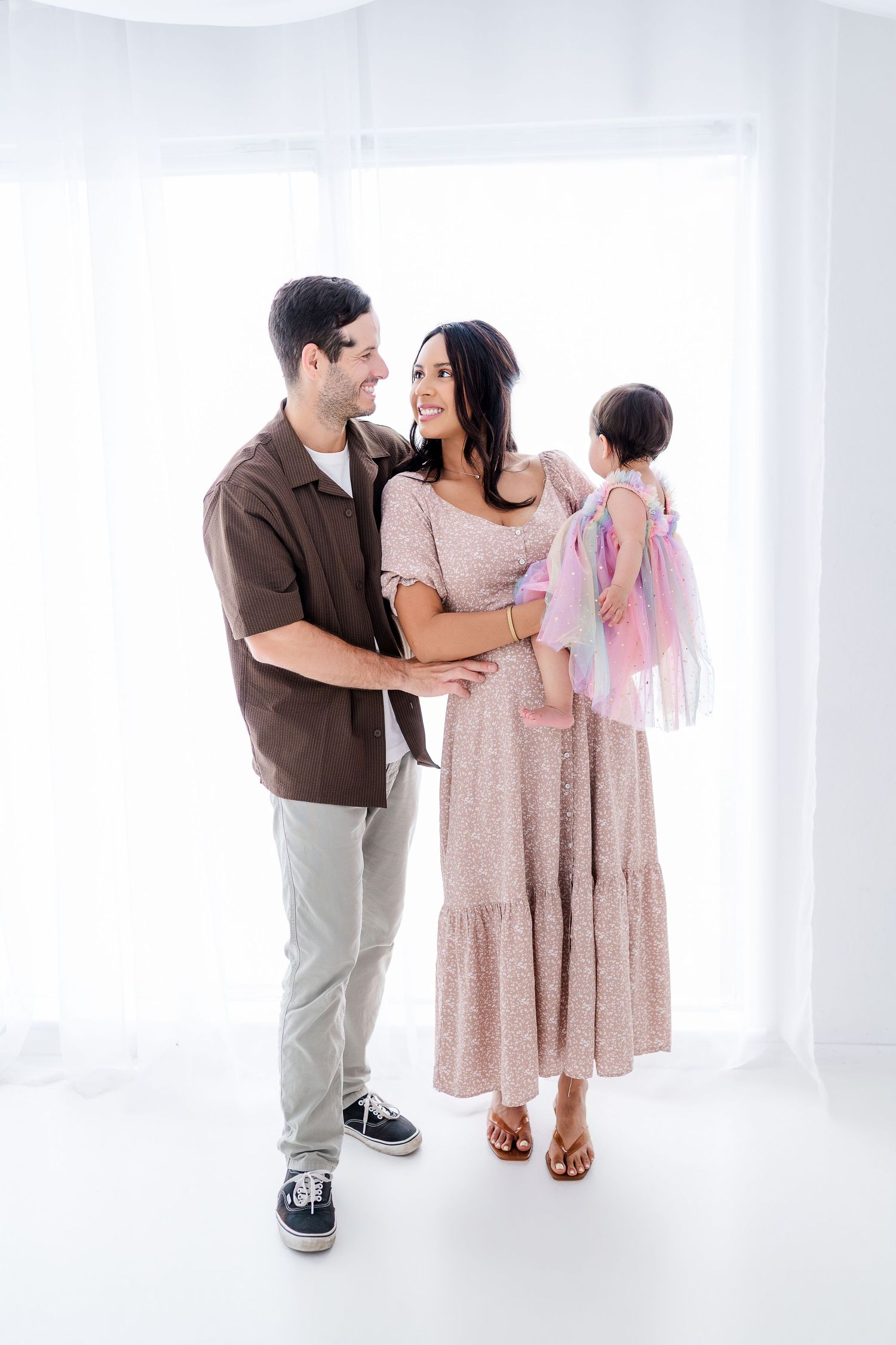 A happy family of three poses together, mom in a floral dress holds a toddler in fairy wings, dad in brown shirt smiles.