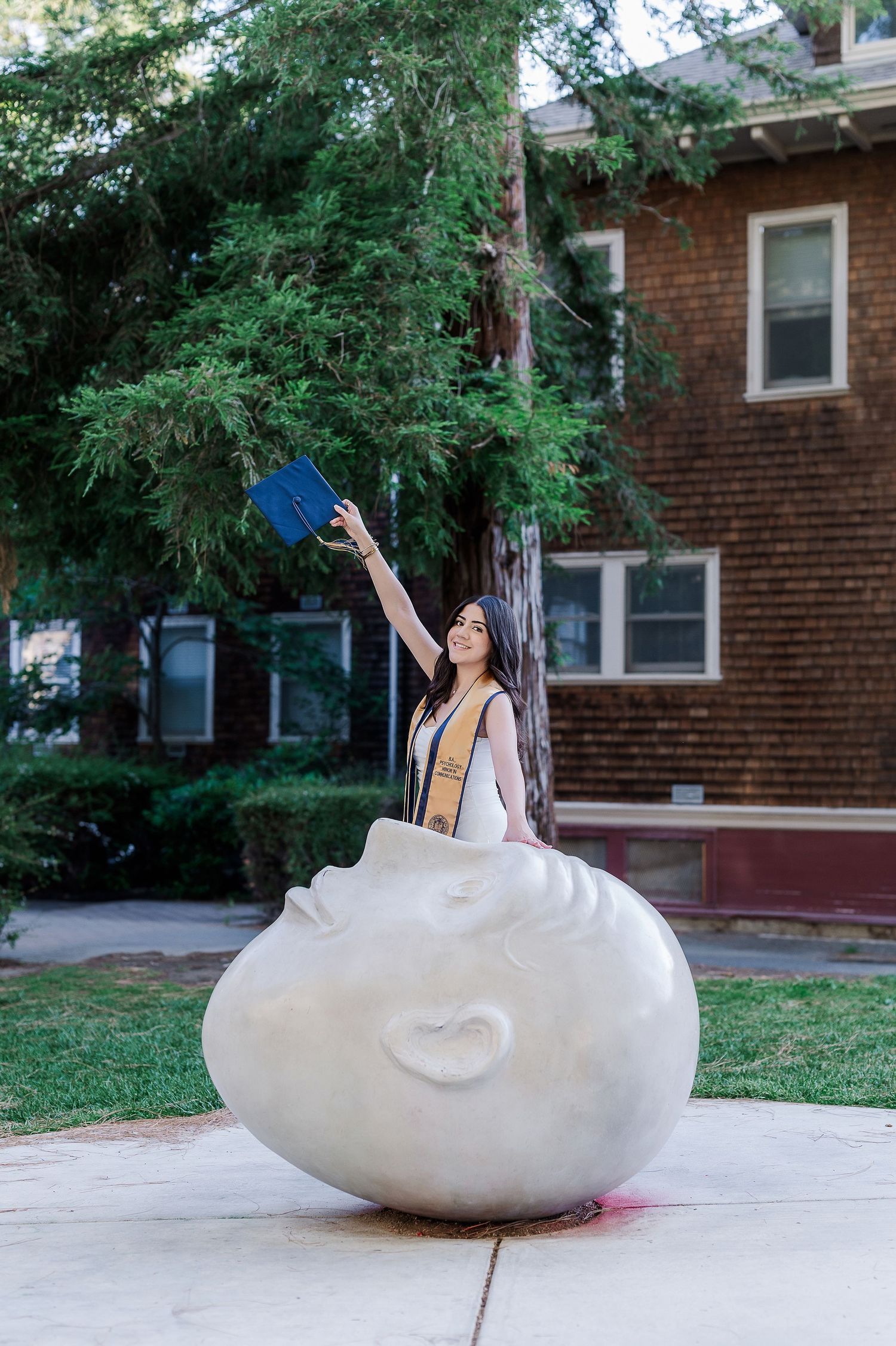 Graduate celebrates on large white egg sculpture on campus grounds.