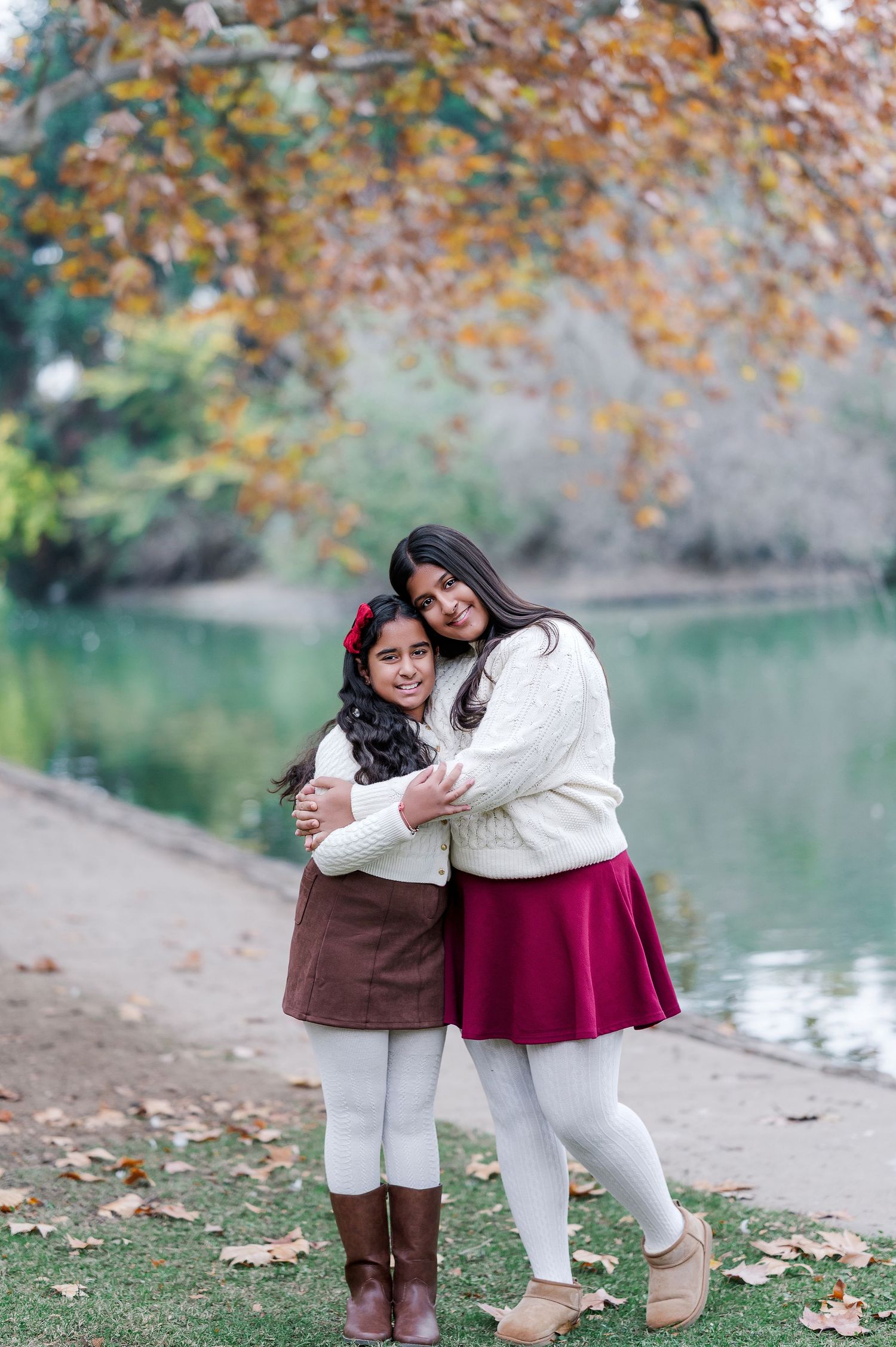 Two people in white and burgundy outfits embrace and laugh together by a lake during autumn.