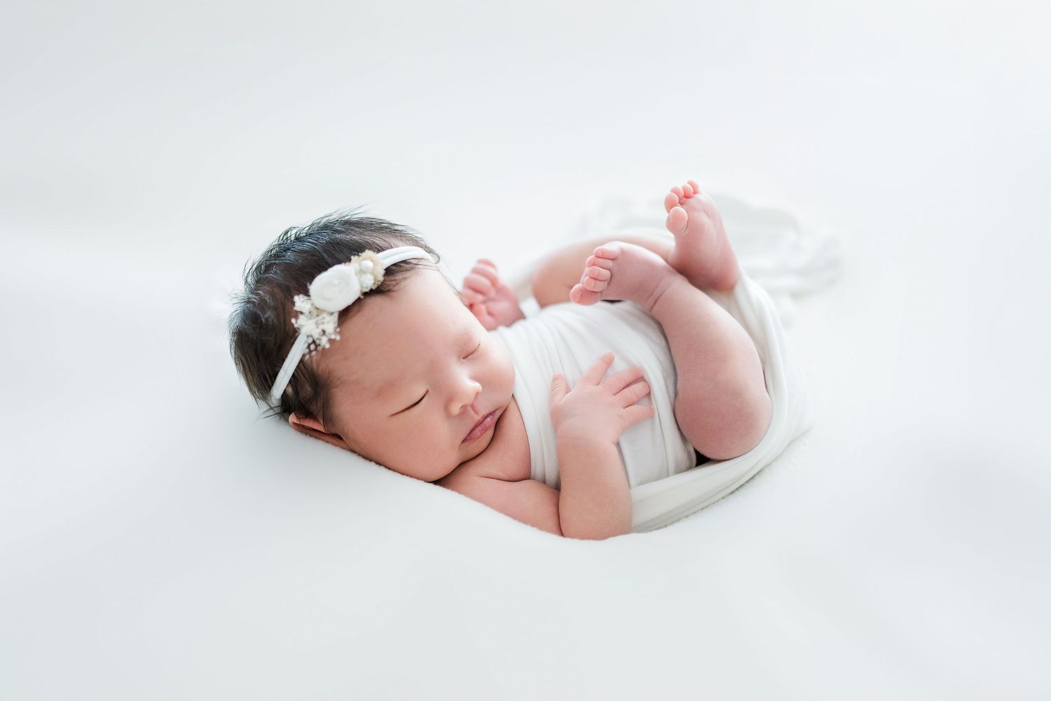Newborn baby wearing white headband sleeping peacefully on white backdrop.